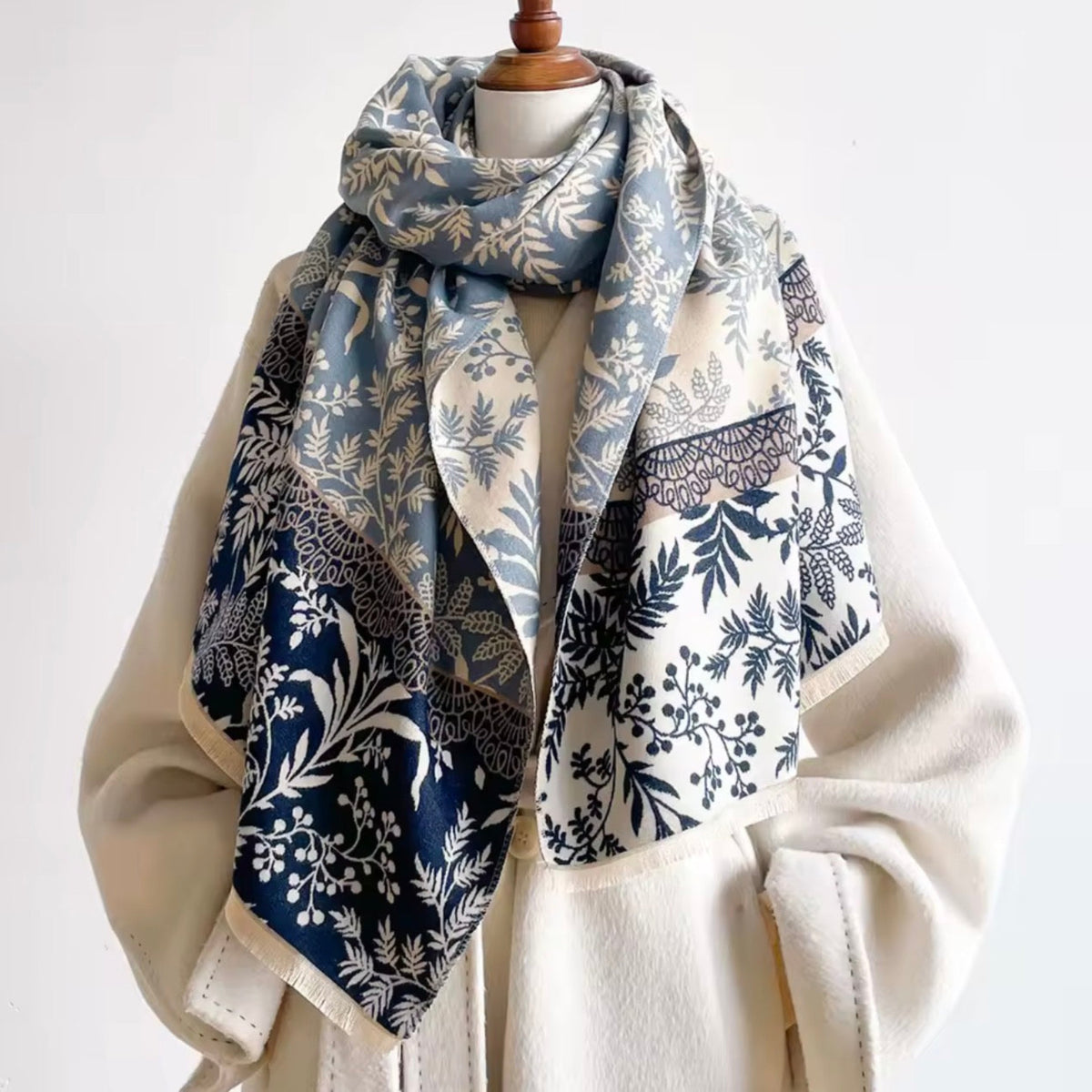 A patterned scarf draped over a cream-colored coat on a mannequin torso against a plain white background. The scarf features botanical and floral motifs in navy blue, dusty blue, and cream.