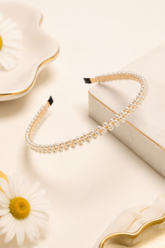 A pearl-studded headband with a gold-toned inner band, styled on a cream-colored surface with daisy flowers and a decorative box.
