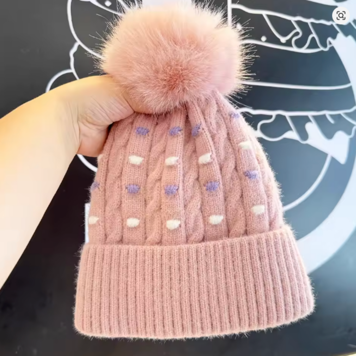 A pink cable-knit beanie hat with a large fluffy pom-pom on top, held by a hand against a dark graphic background.