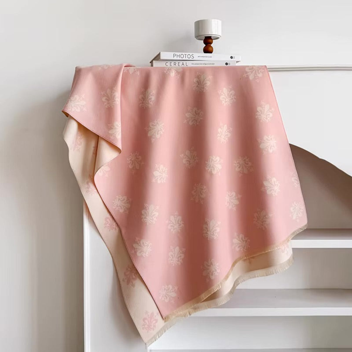 A pink fabric throw blanket with a cream floral pattern and fringed edges, draped over a white shelf in a minimalist room setting.