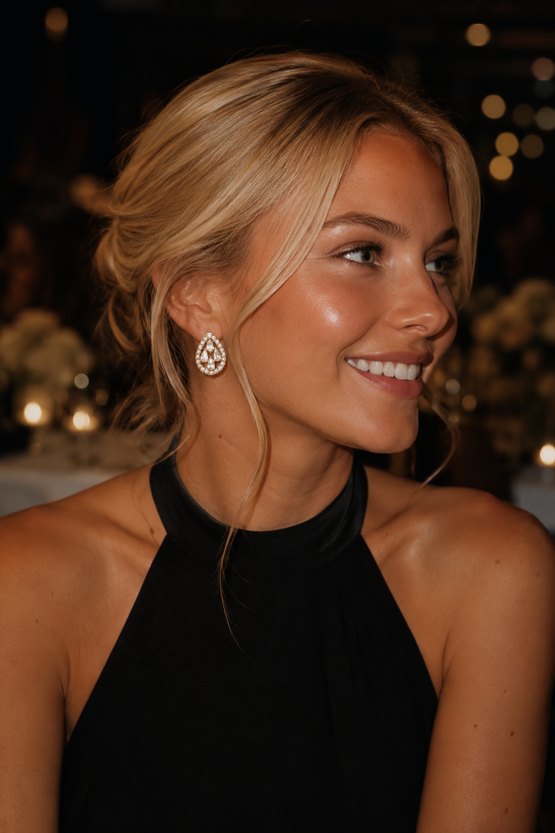 A smiling blonde woman in a black halter top wearing a teardrop-shaped diamond stud earring, photographed in a dimly lit event setting with warm bokeh lights in the background.