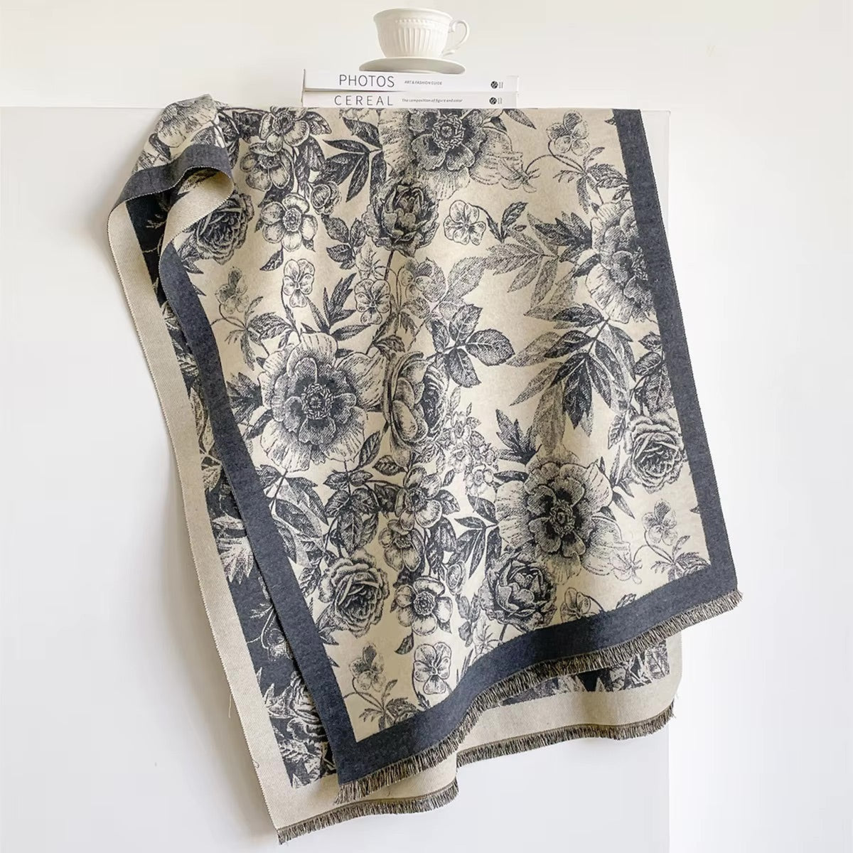 A square scarf with a black floral toile pattern on a cream background, featuring a dark navy border and fringe, draped over a white display stand.