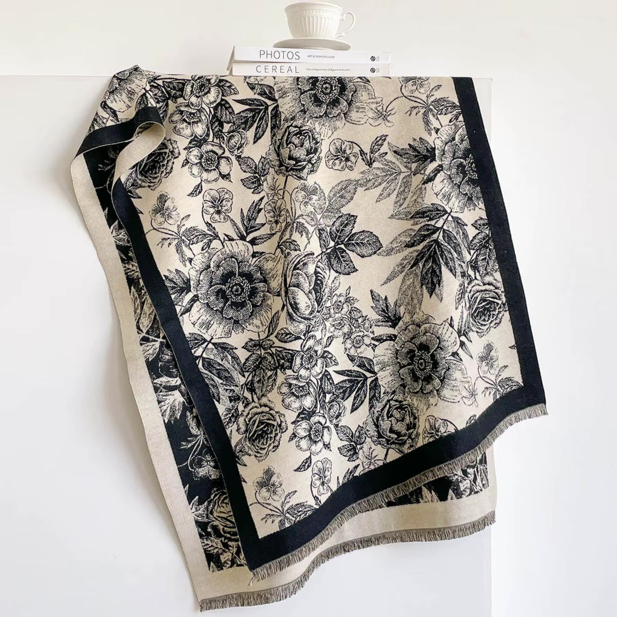 A square scarf with a black floral toile pattern on a cream background, featuring a solid black border and fringe, draped over a white surface with a book and cup in the background.