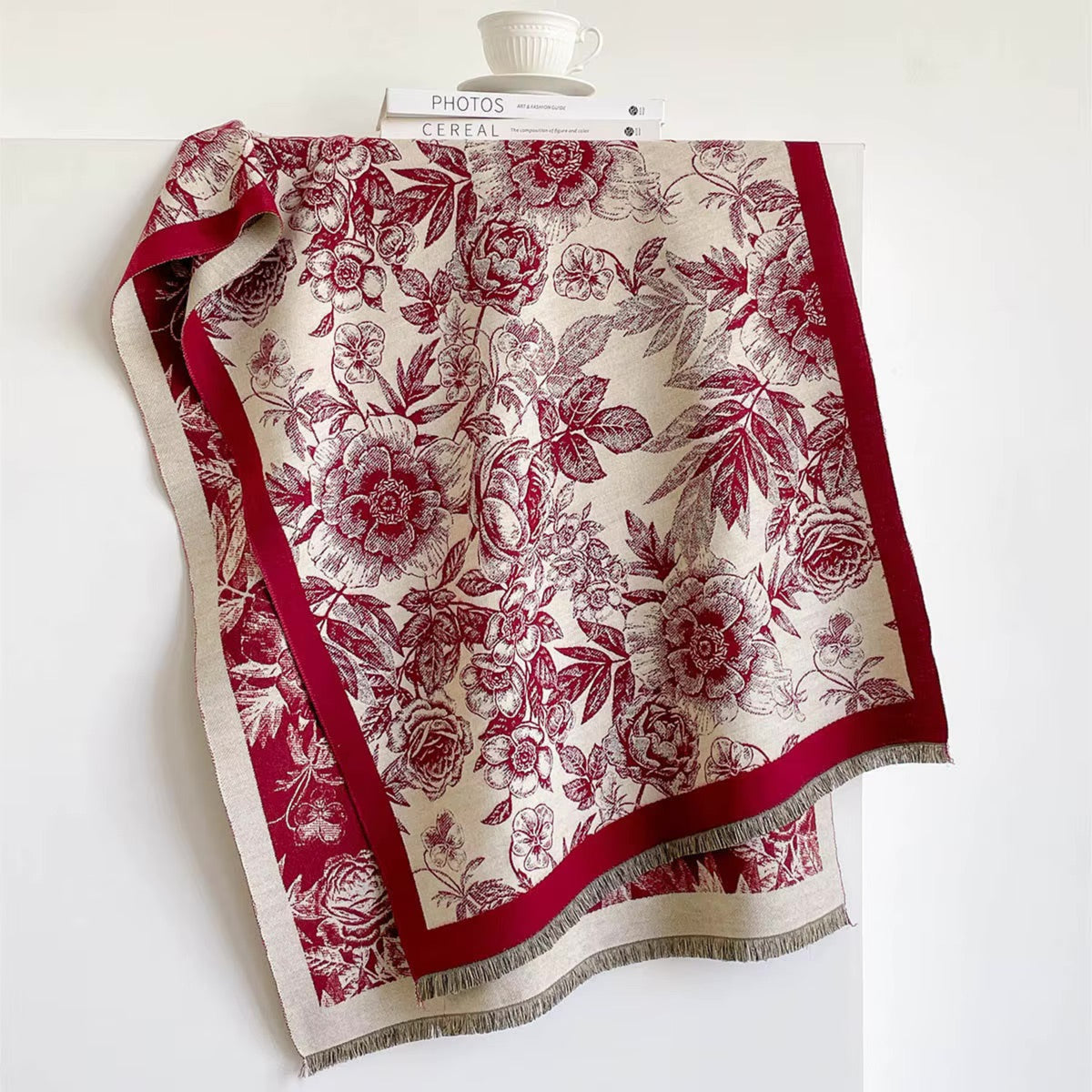A square scarf with a classic toile-style floral print in deep red and beige, draped over a white surface with a book and cup visible at the top.