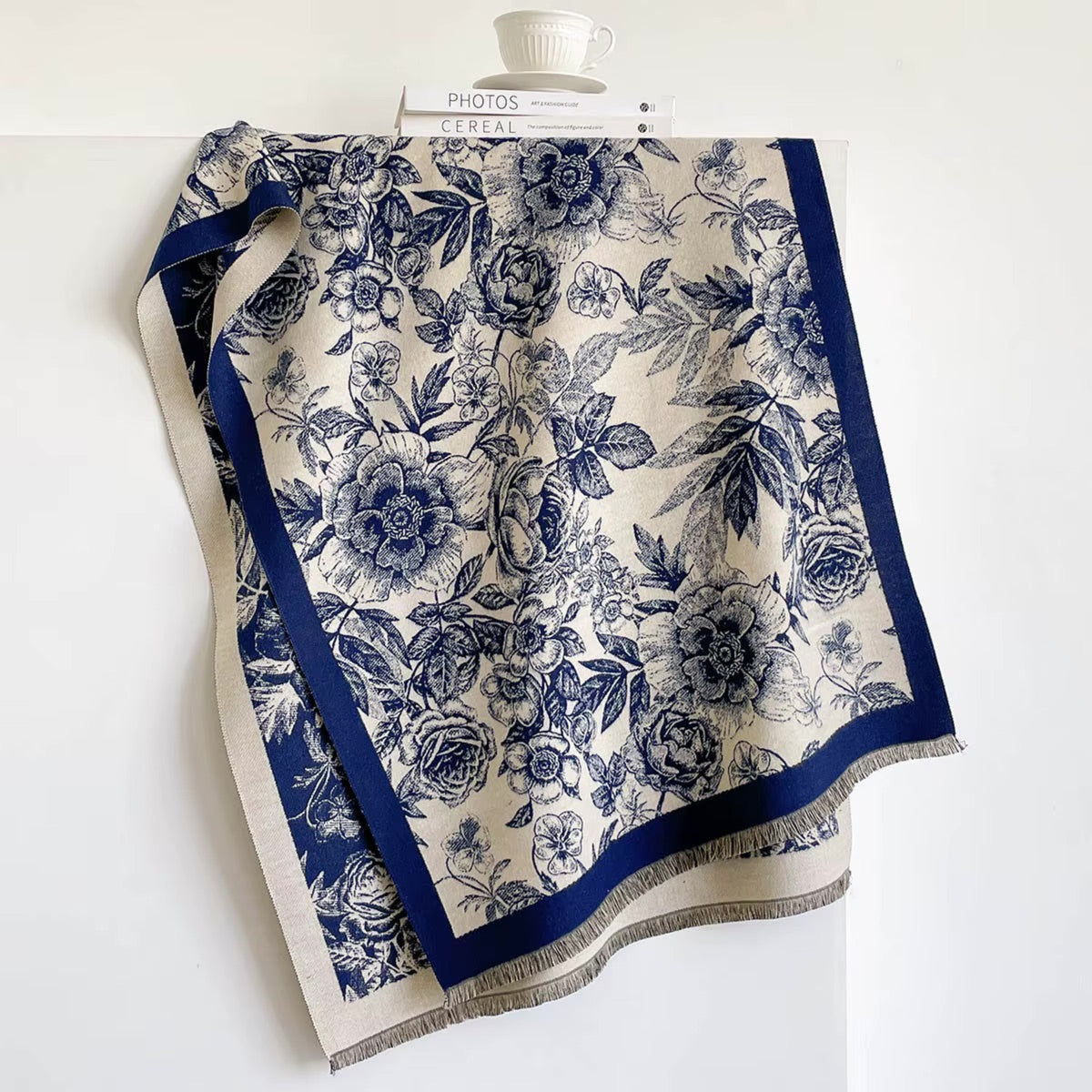 A square scarf with a navy blue floral toile pattern on a cream background, featuring a solid navy border and fringed edges, draped over a white display stand.