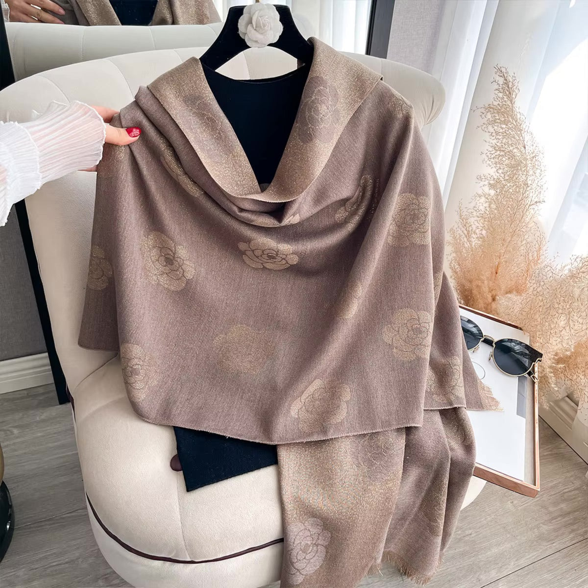 A taupe-brown shawl with a subtle tonal floral jacquard pattern draped over a black top on a hanger, styled on a cream-colored armchair with a hand holding its edge.