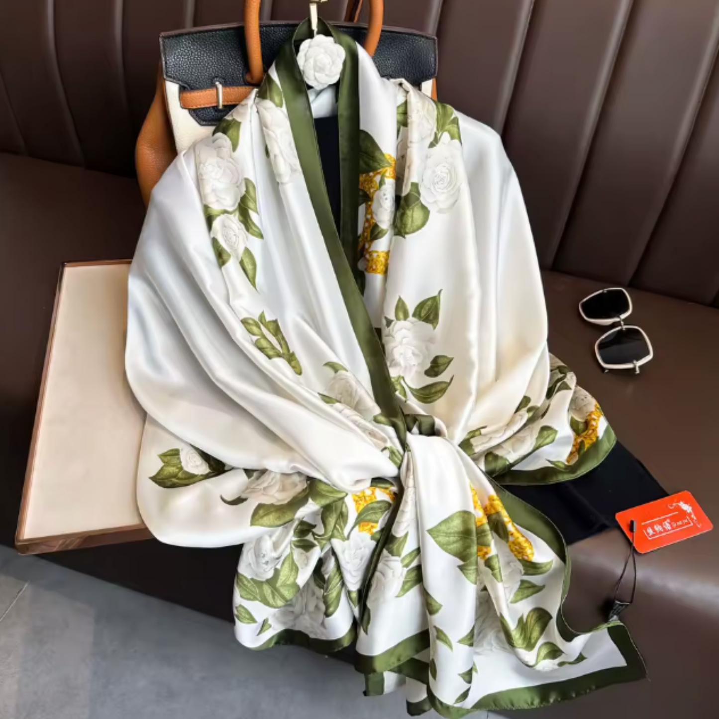 A white silk scarf with a green and yellow botanical print draped over a brown leather handbag on a brown leather bench. A small red tag is attached to the scarf.