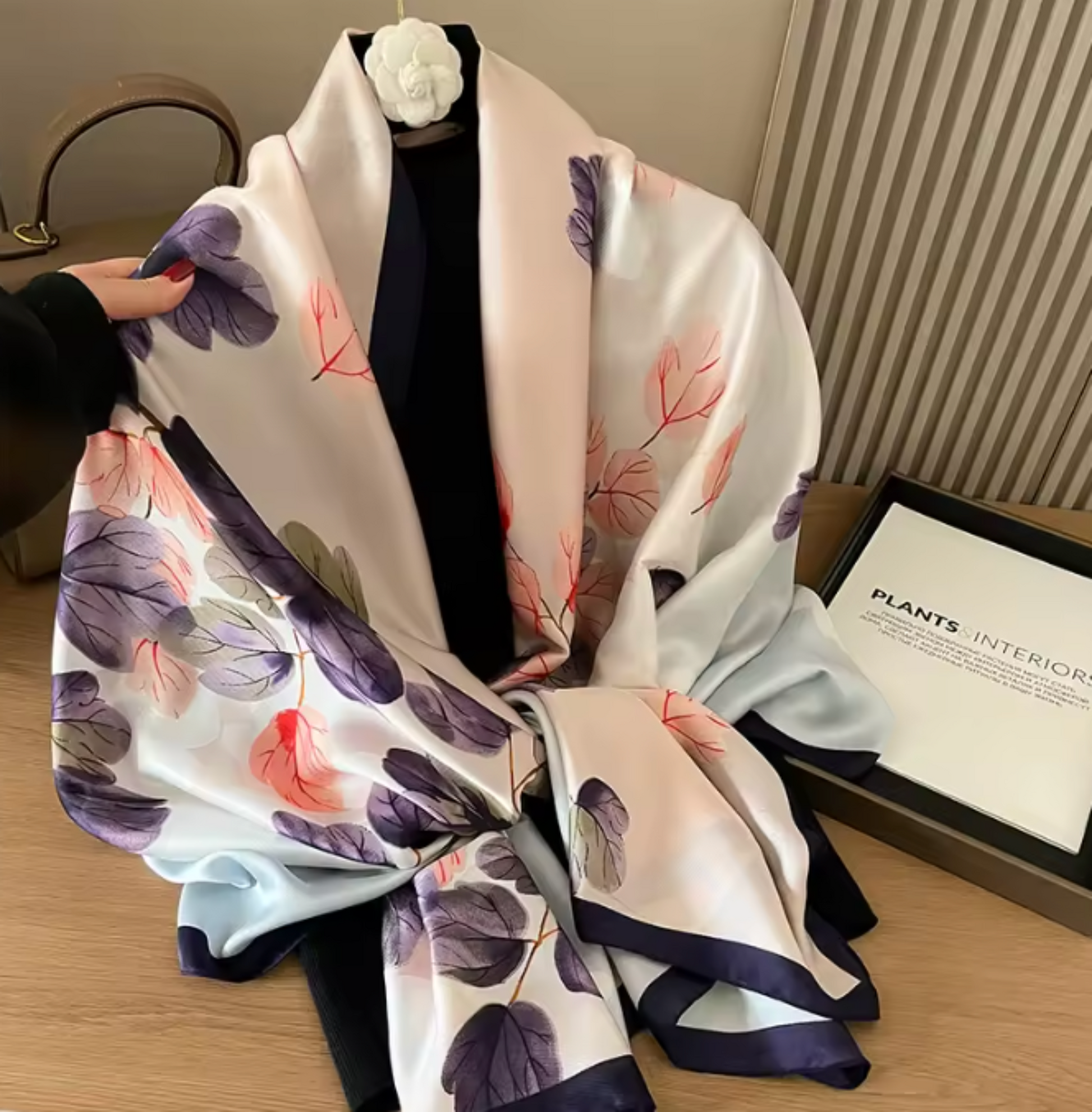 A white silk scarf with a purple and pink botanical leaf pattern and dark navy borders, draped over a black dress form decorated with a white camellia flower. A hand is holding the left side of the scarf, and a framed print reading 'PLANTS INTERIOR' sits on a wooden table next to it.