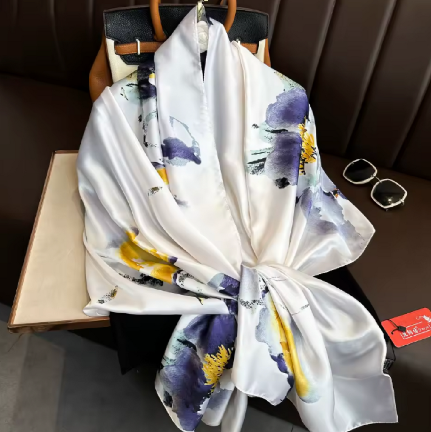 A white silk scarf with a watercolor-style print in purple, yellow, and gray, draped over a black handbag on a dark brown leather bench.