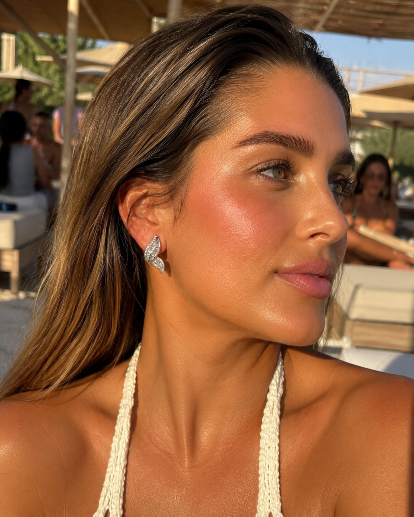 A woman in a beach resort setting wearing a silver leaf-shaped pavé stud earring, with warm golden hour sunlight illuminating her face and the background.