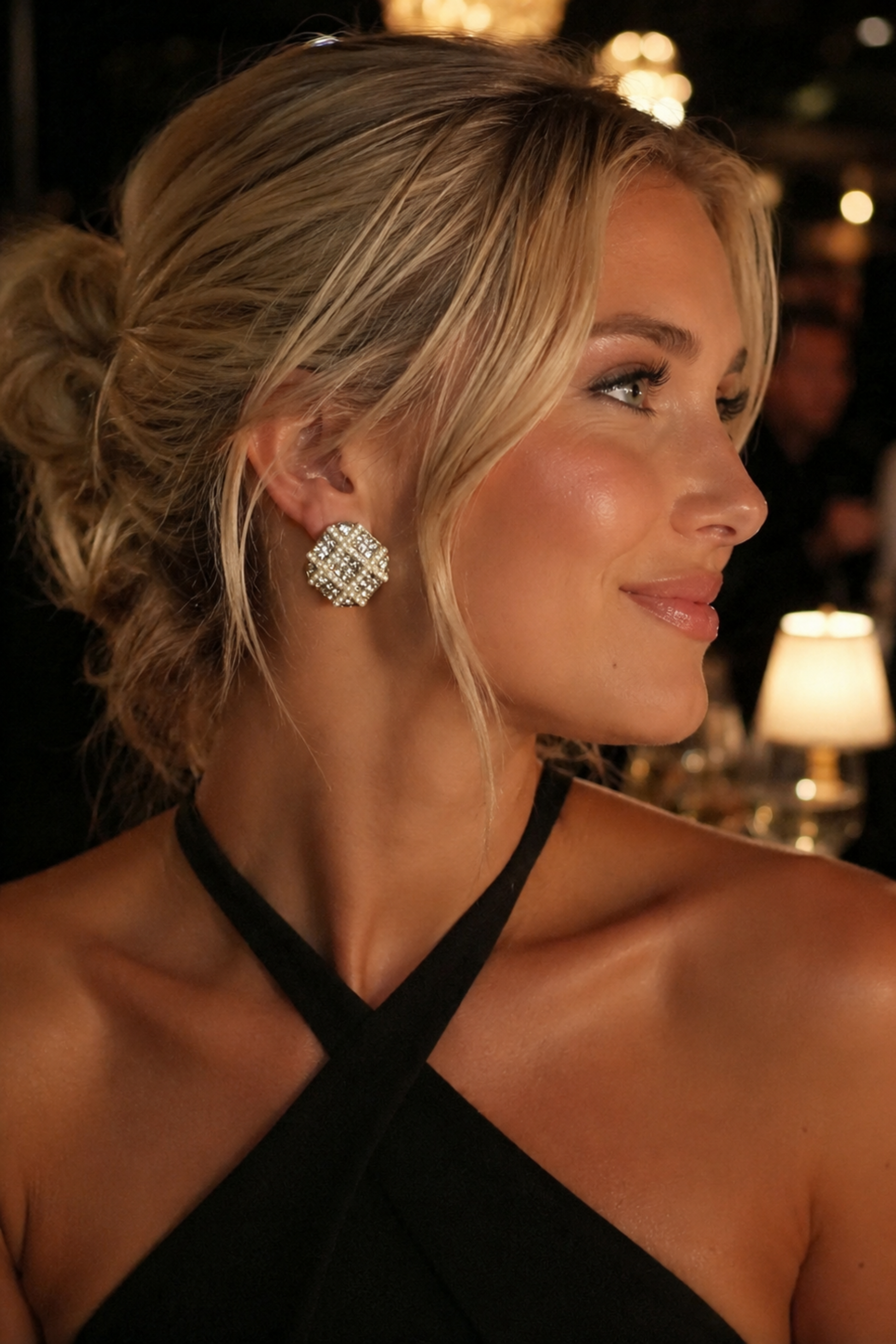 A woman in a black halter dress wearing a large, sparkling silver geometric stud earring, photographed in a dimly lit restaurant setting.