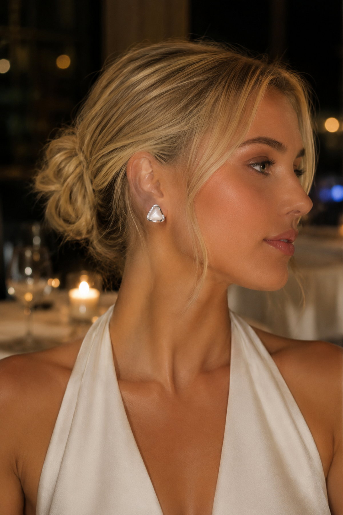 A woman in a white halter dress wearing a small white triangular stud earring, photographed in a softly lit indoor setting with blurred lights in the background.