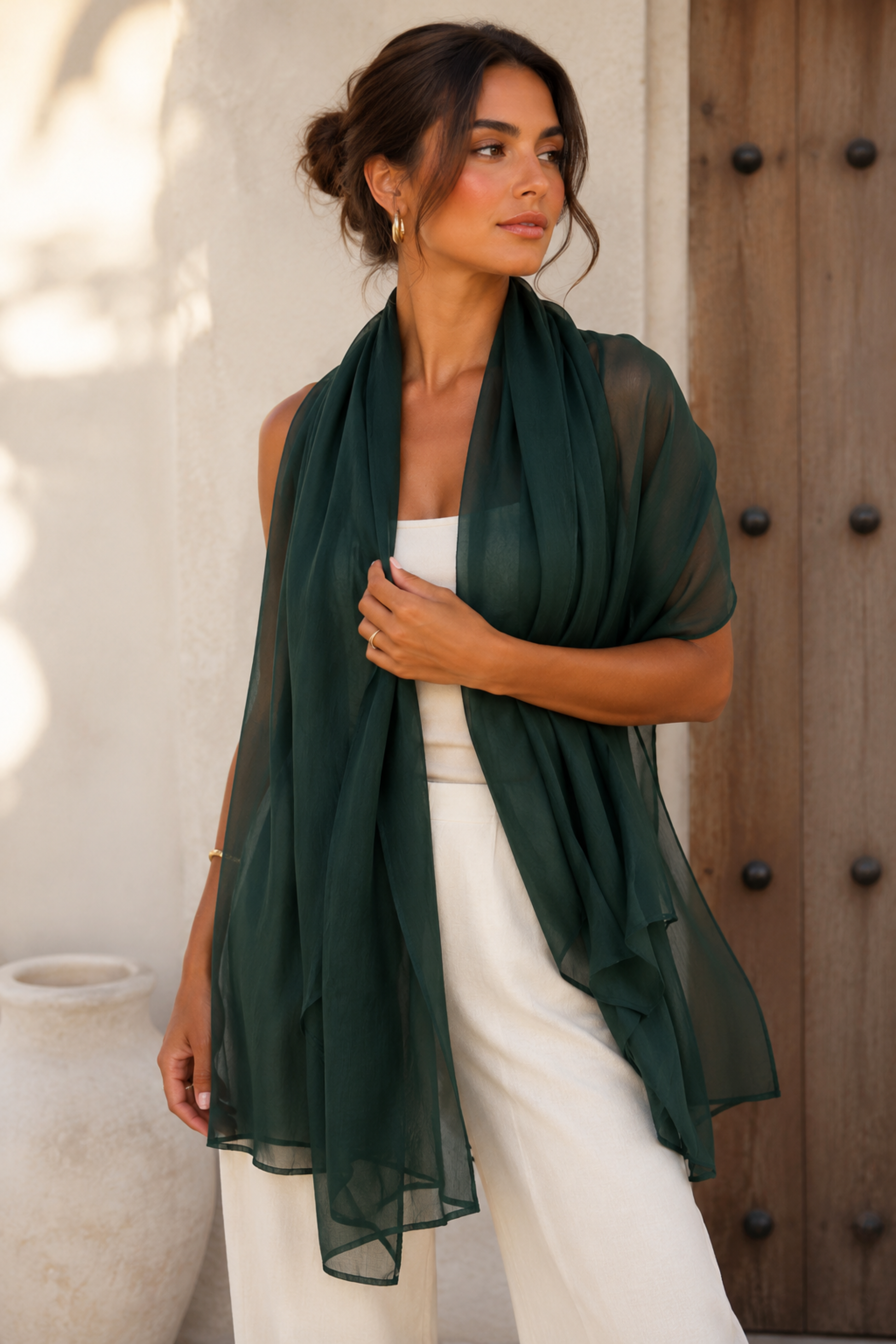 A woman models a dark forest green sheer chiffon shawl draped over her shoulders, paired with a white top and white pants, standing in front of a rustic wooden door and white stucco wall.