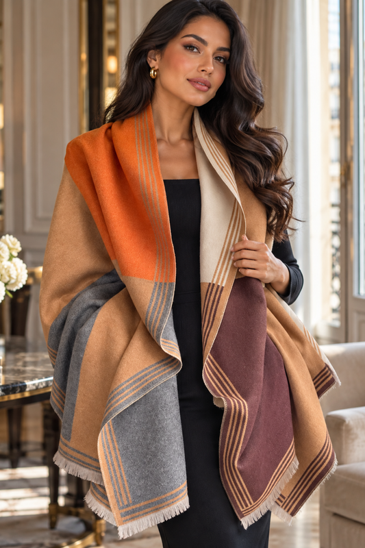 A woman models a large, multi-colored fringed scarf or shawl in an elegant indoor setting. The scarf features large color-blocked sections of orange, beige, gray, and burgundy with striped borders.