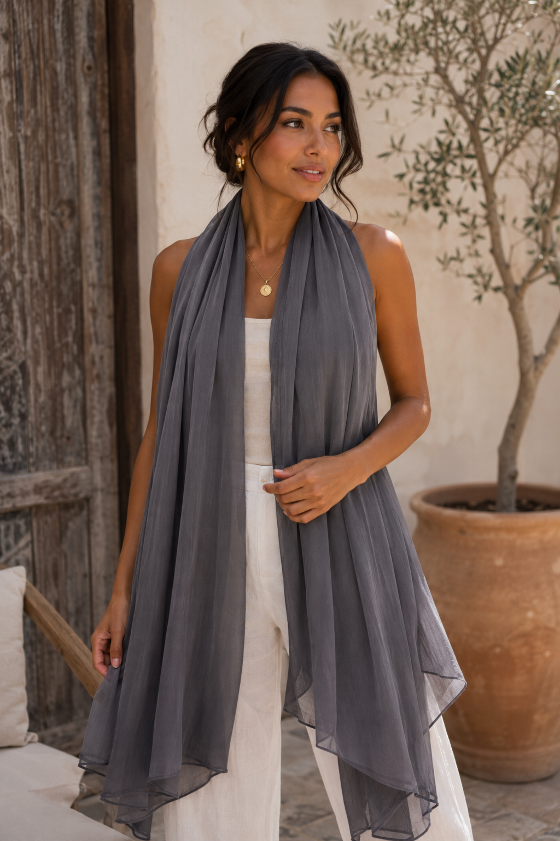 A woman models a lightweight, semi-sheer gray scarf draped over her shoulders, paired with a white sleeveless top and white trousers in an outdoor rustic setting with a potted olive tree.