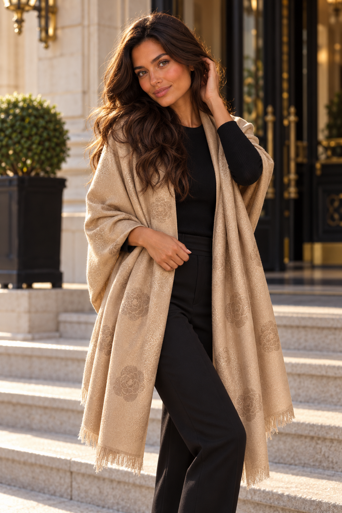 A woman stands on outdoor stone steps wearing a beige shawl with tonal circular motifs over a black outfit. The background shows a grand building entrance with gold details and potted plants.