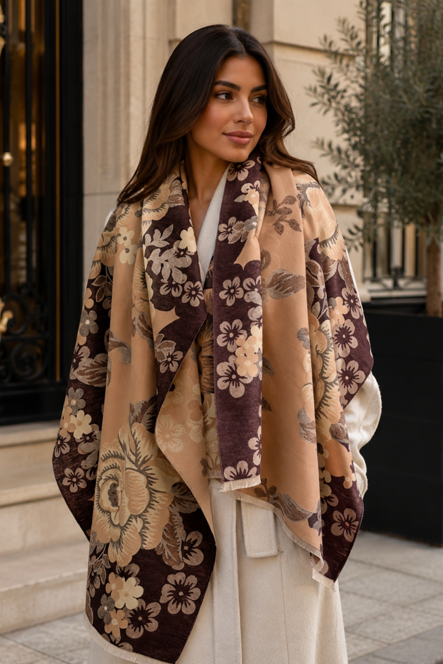 A woman wearing a beige and dark brown floral shawl over a white dress, standing outdoors in front of a building entrance.