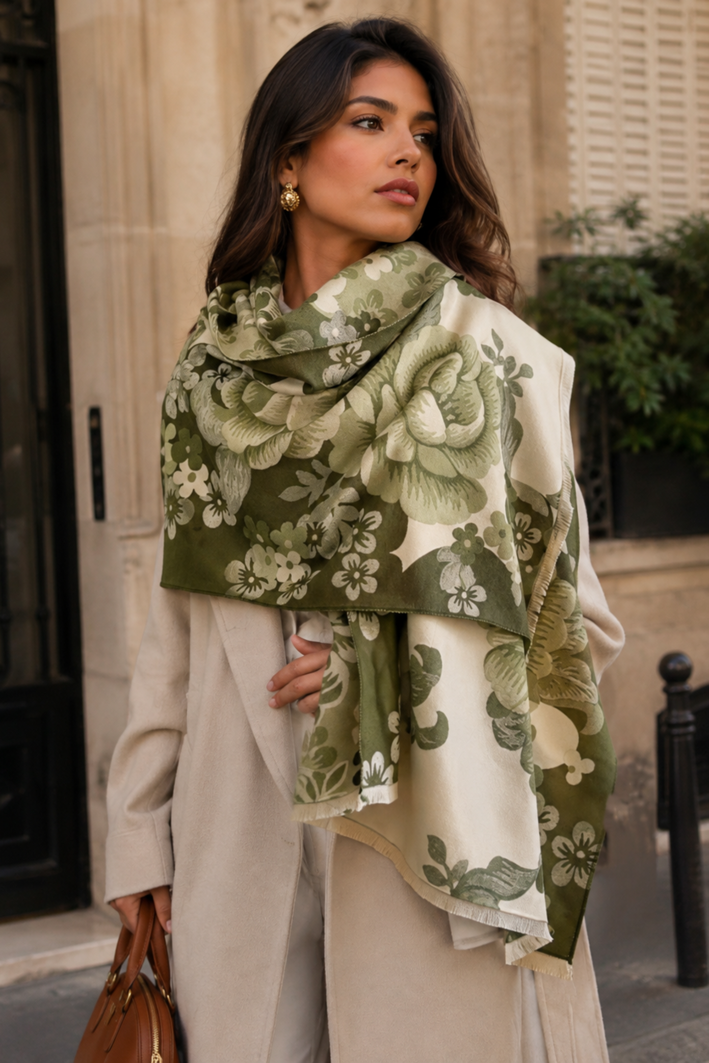 A woman wearing a beige coat and a large floral scarf in olive green and cream, standing outdoors in front of a stone building.