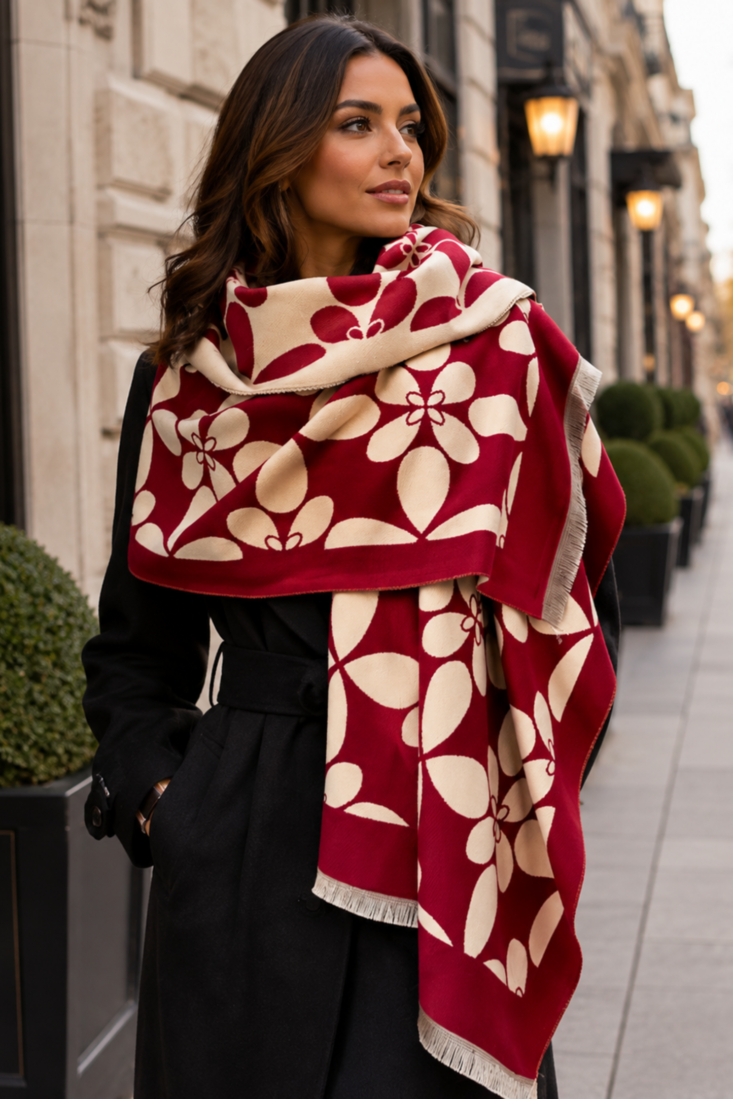 A woman wearing a black coat and a large burgundy and beige floral-patterned scarf, standing on a city sidewalk.