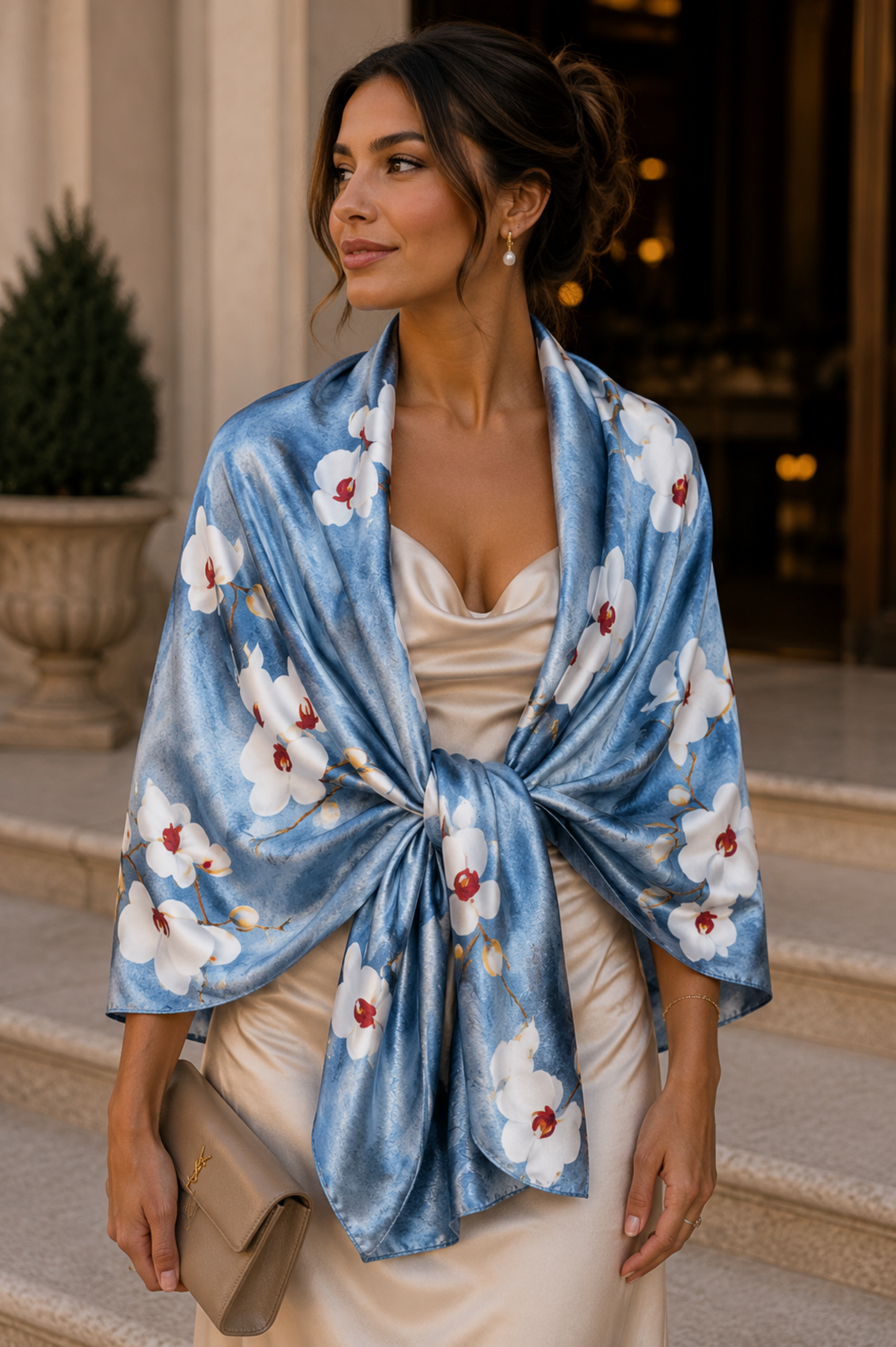 A woman wearing a blue silk-like shawl with a white floral print and red flower centers, tied at the waist over a cream satin dress, holding a taupe clutch on stone steps.