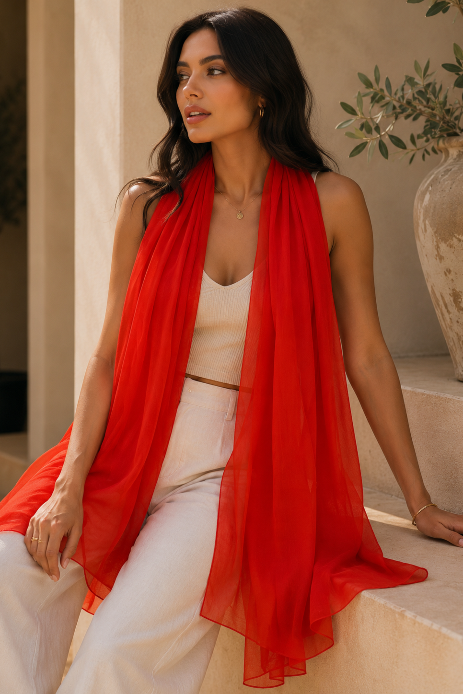 A woman wearing a bright red sheer scarf draped over her shoulders, styled with a white crop top and white wide-leg pants, sitting on stone steps in a warm, sunlit setting.