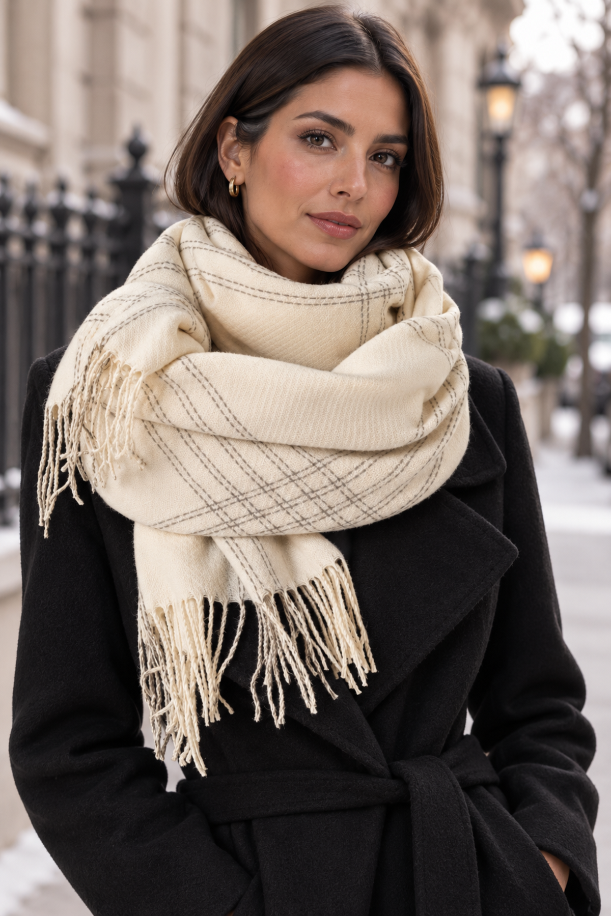 A woman wearing a cream-colored plaid scarf with fringe trim and a black coat, photographed outdoors on a city street.