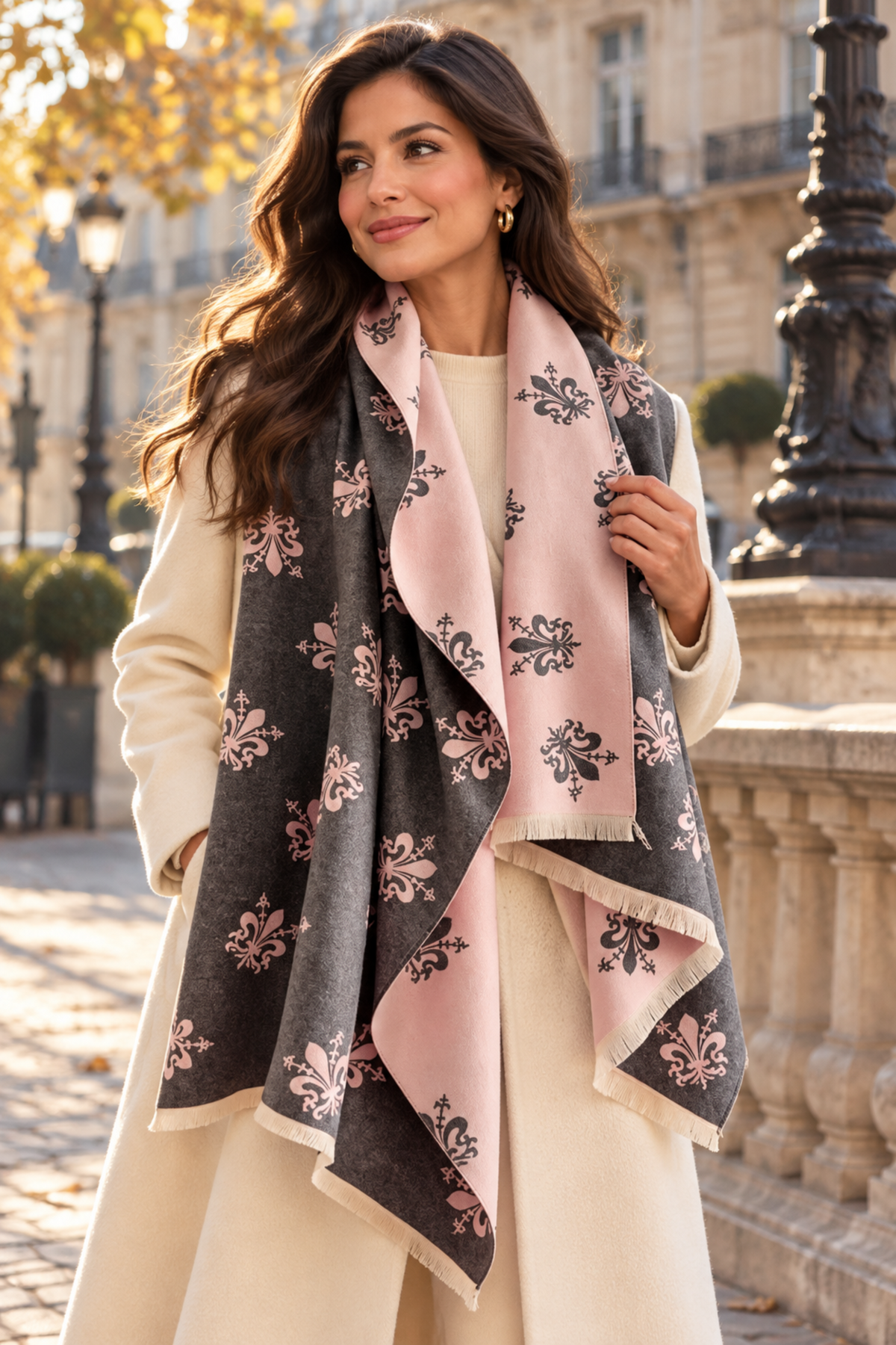 A woman wearing a cream coat and a large patterned scarf in dusty pink and charcoal gray with black floral motifs, standing outdoors in front of a stone building.
