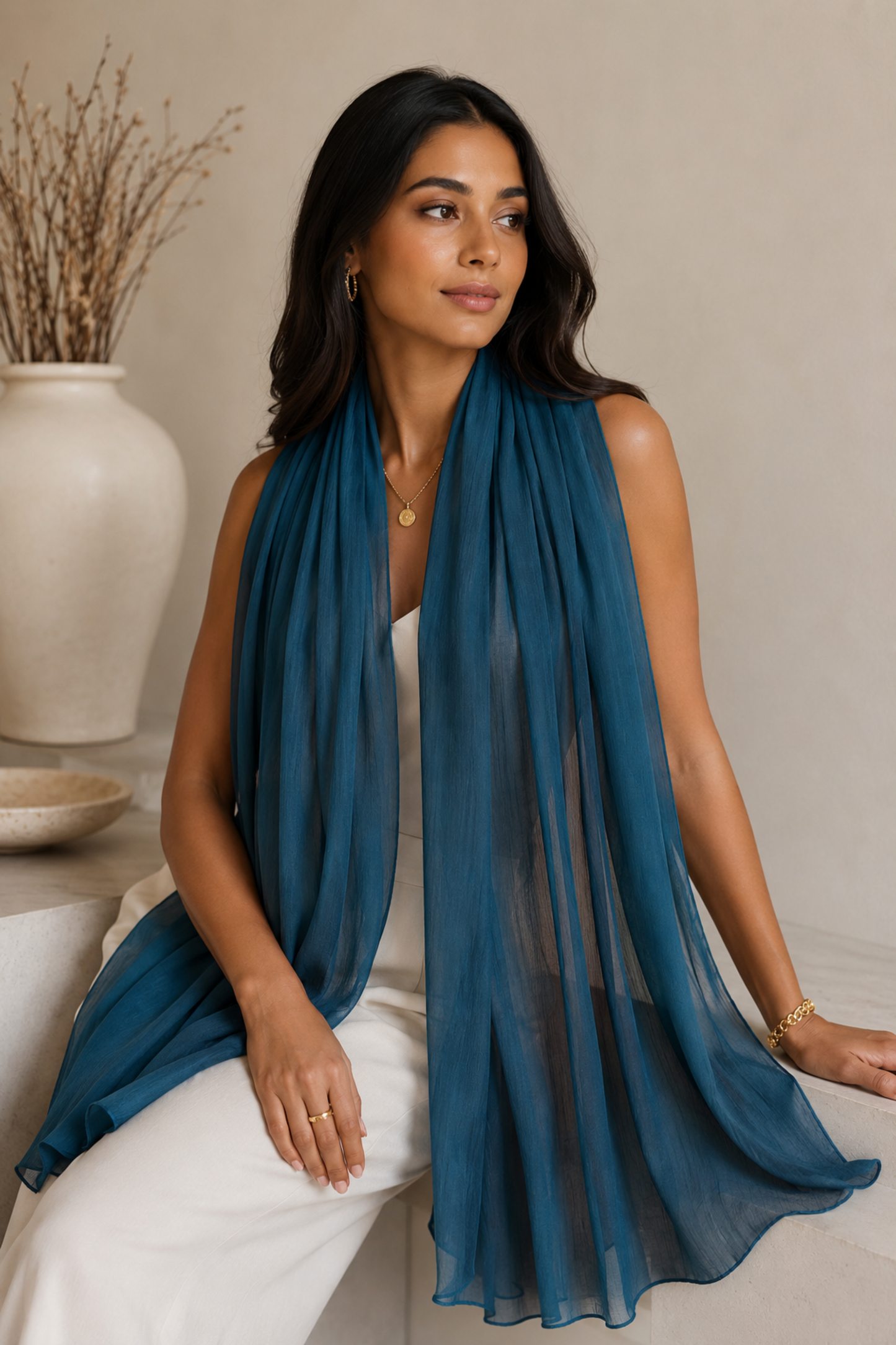 A woman wearing a flowing teal blue sheer scarf draped over her shoulders, paired with white pants, posed in a warm, neutral-toned indoor setting.
