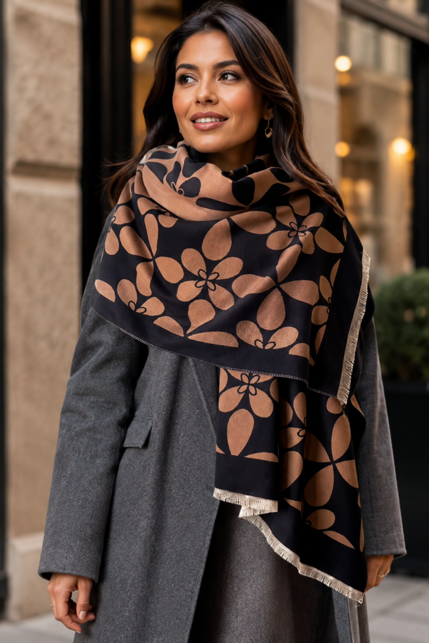 A woman wearing a gray coat and a black scarf with a large tan floral pattern, standing outdoors in front of a city building.