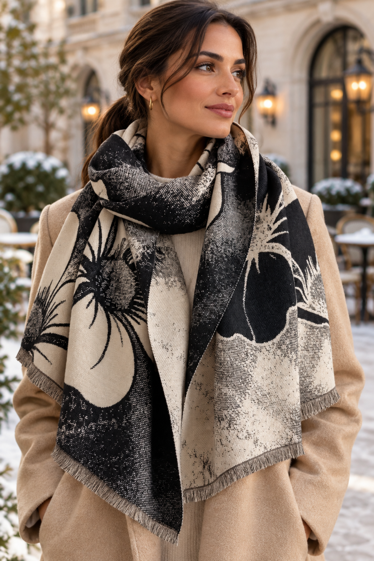 A woman wearing a large black and cream floral scarf with fringed edges over a beige coat, photographed outdoors in front of a softly blurred building facade.