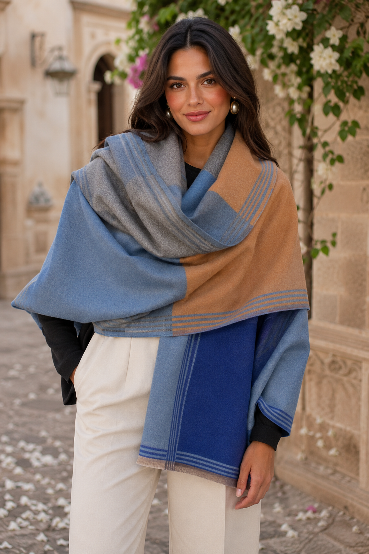 A woman wearing a large color-blocked knit shawl wrap in shades of dusty blue, camel tan, and royal blue, styled with white trousers and a black top, standing outdoors in front of a stone building.