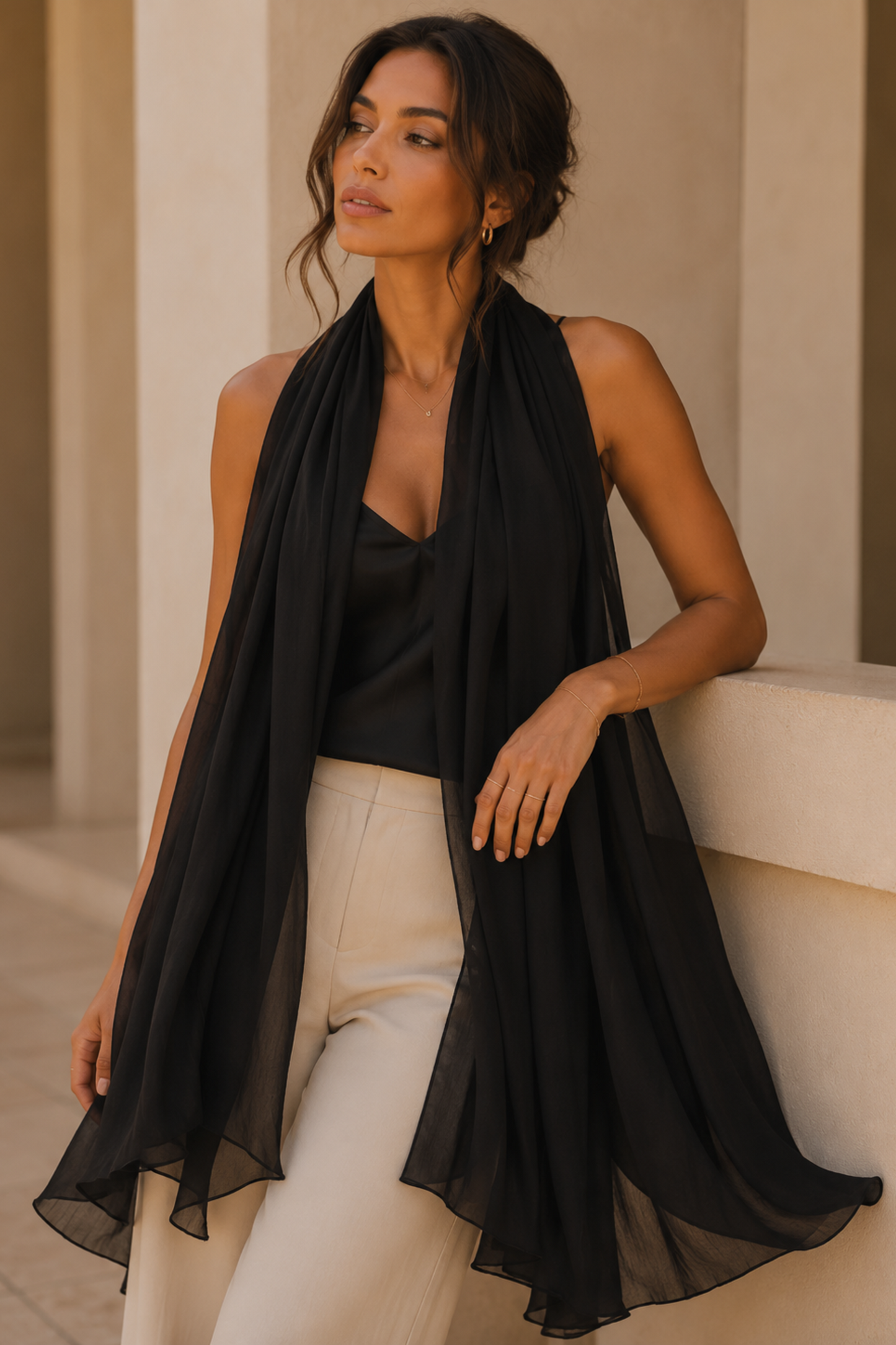 A woman wearing a sheer black chiffon wrap scarf draped over a black camisole top and cream-colored trousers, leaning against a beige stone wall.