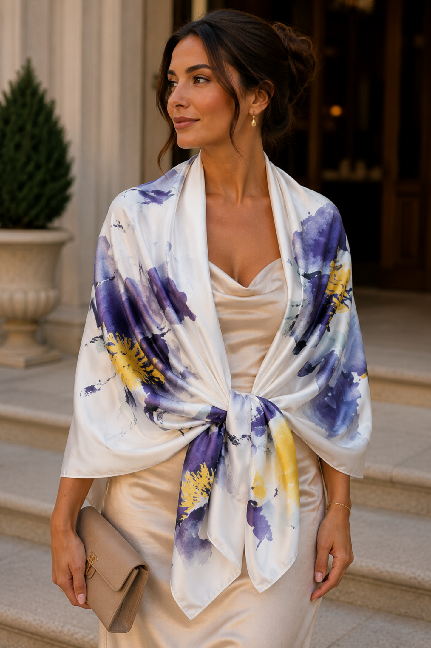 A woman wearing a white silk shawl with purple and yellow watercolor floral patterns tied around her shoulders, styled over a cream satin dress while holding a beige clutch on stone steps.