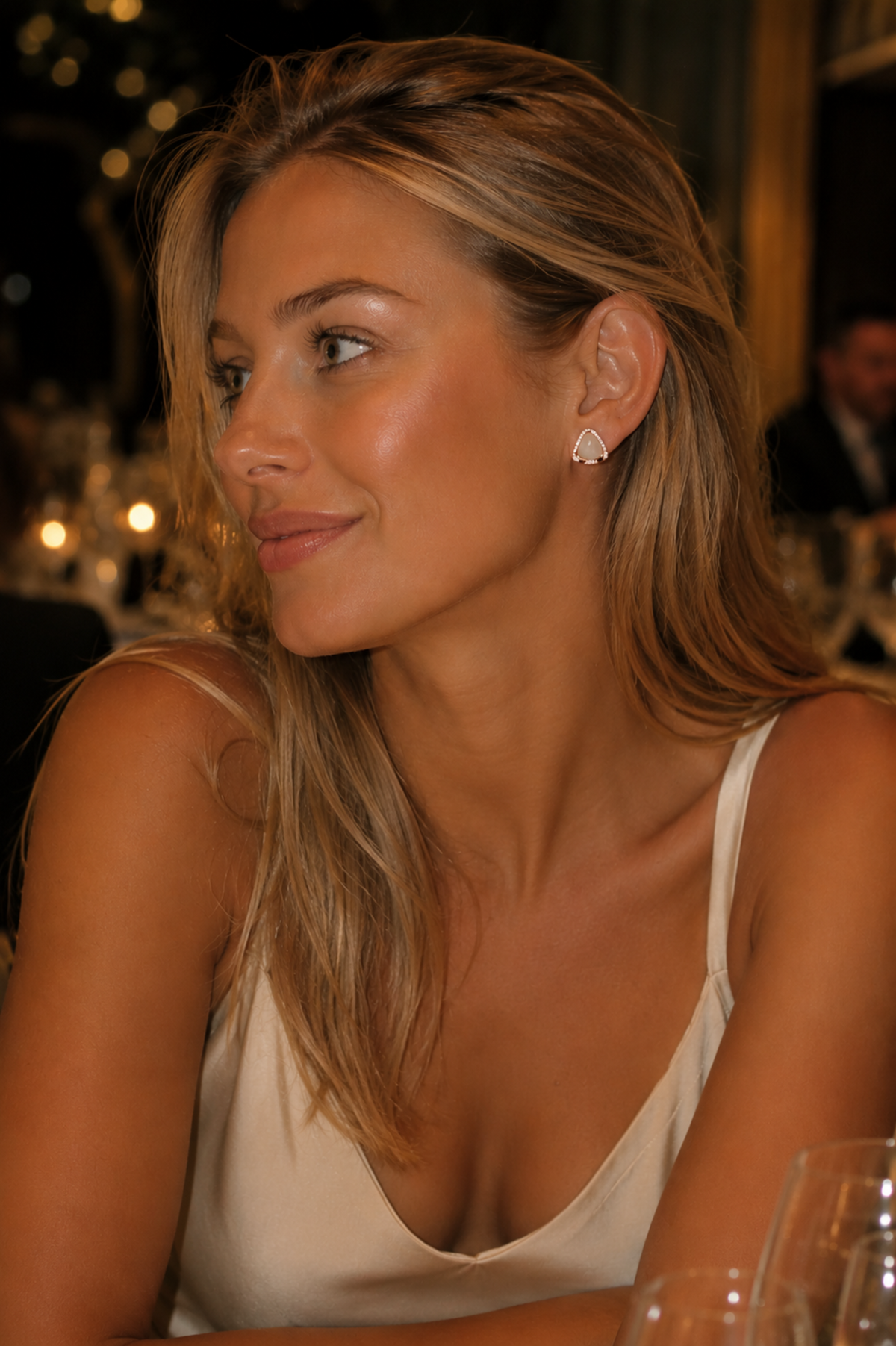 A woman with blonde hair wearing a white spaghetti-strap top and a small silver hoop earring, photographed in a dimly lit indoor setting.