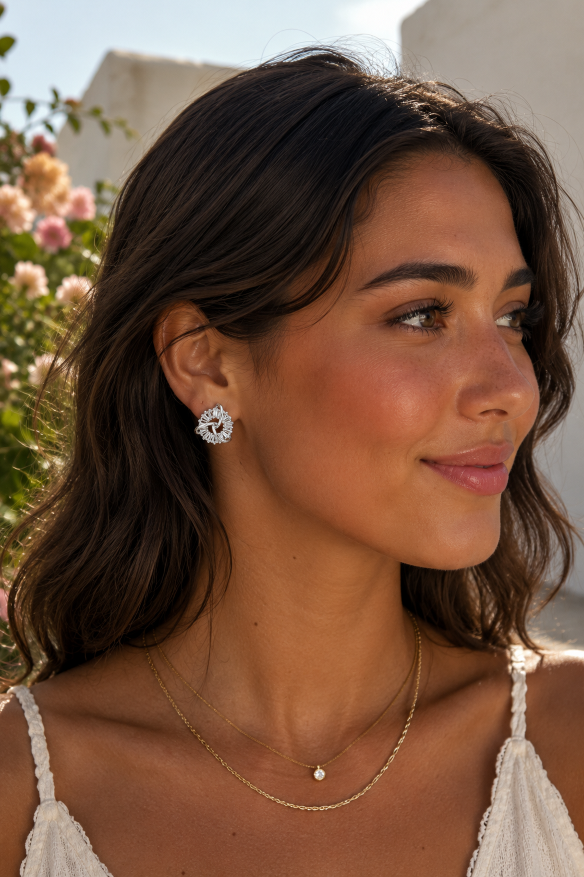 A woman with dark brown hair is shown in a close-up profile, wearing a silver-toned floral stud earring, a delicate gold chain necklace with a small white bead, and a second gold chain necklace.