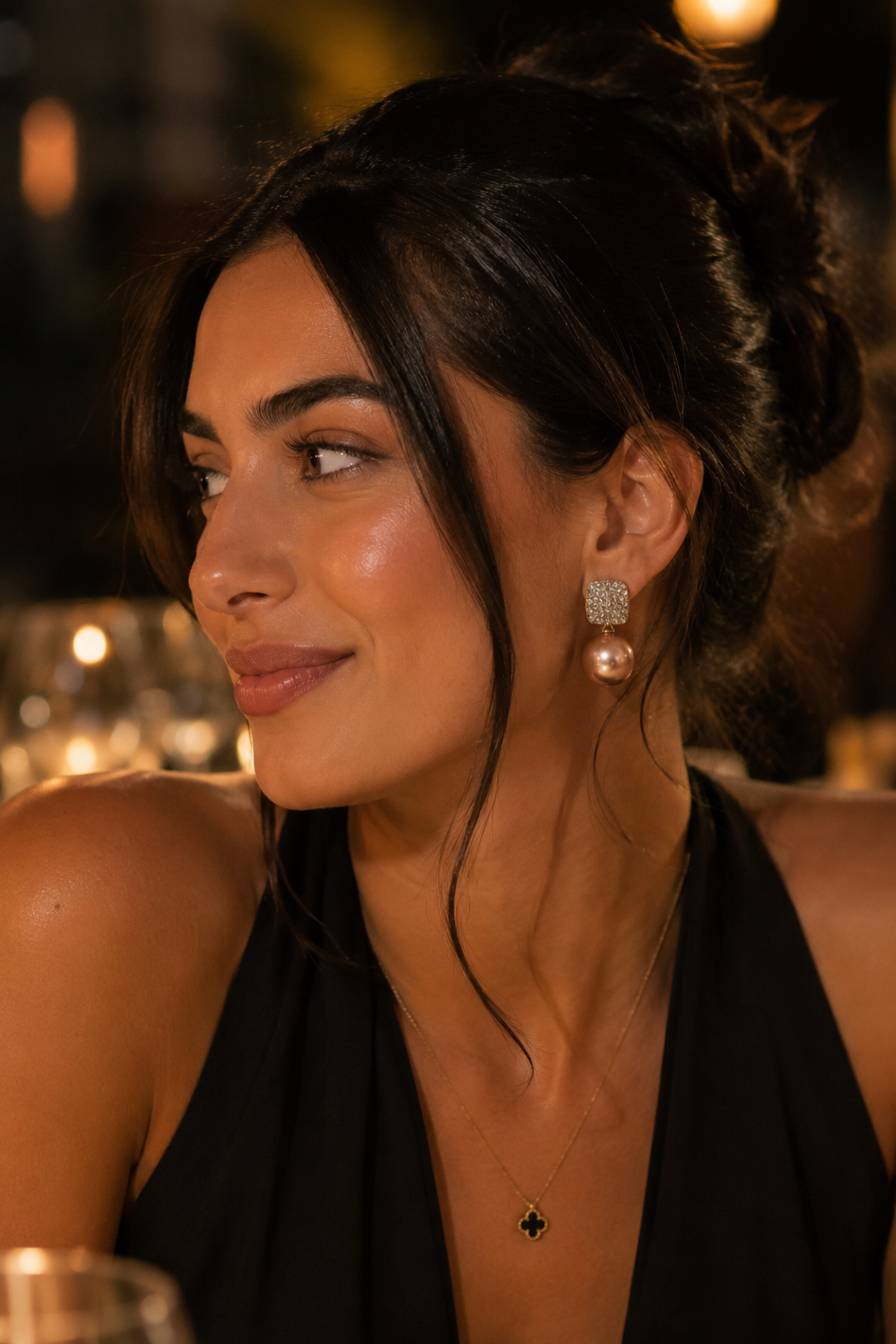 A woman with dark hair in a half-up bun wearing a black halter top, a small black cross pendant necklace, and a textured silver and pearl drop earring, captured in a warm, dimly lit social setting.