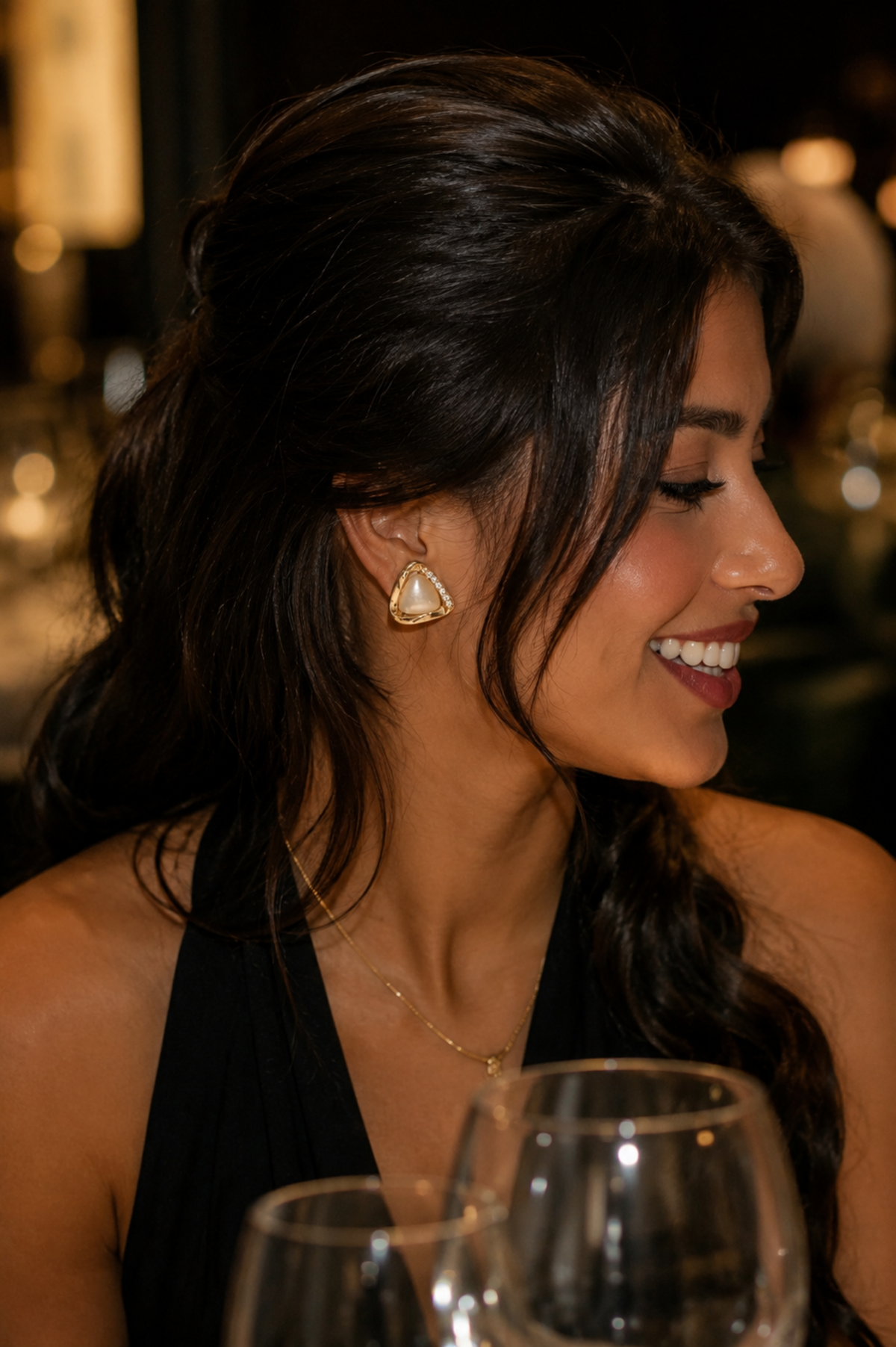 A woman with dark hair wearing a gold triangular stud earring, smiling in a dimly lit indoor setting with wine glasses in the foreground.