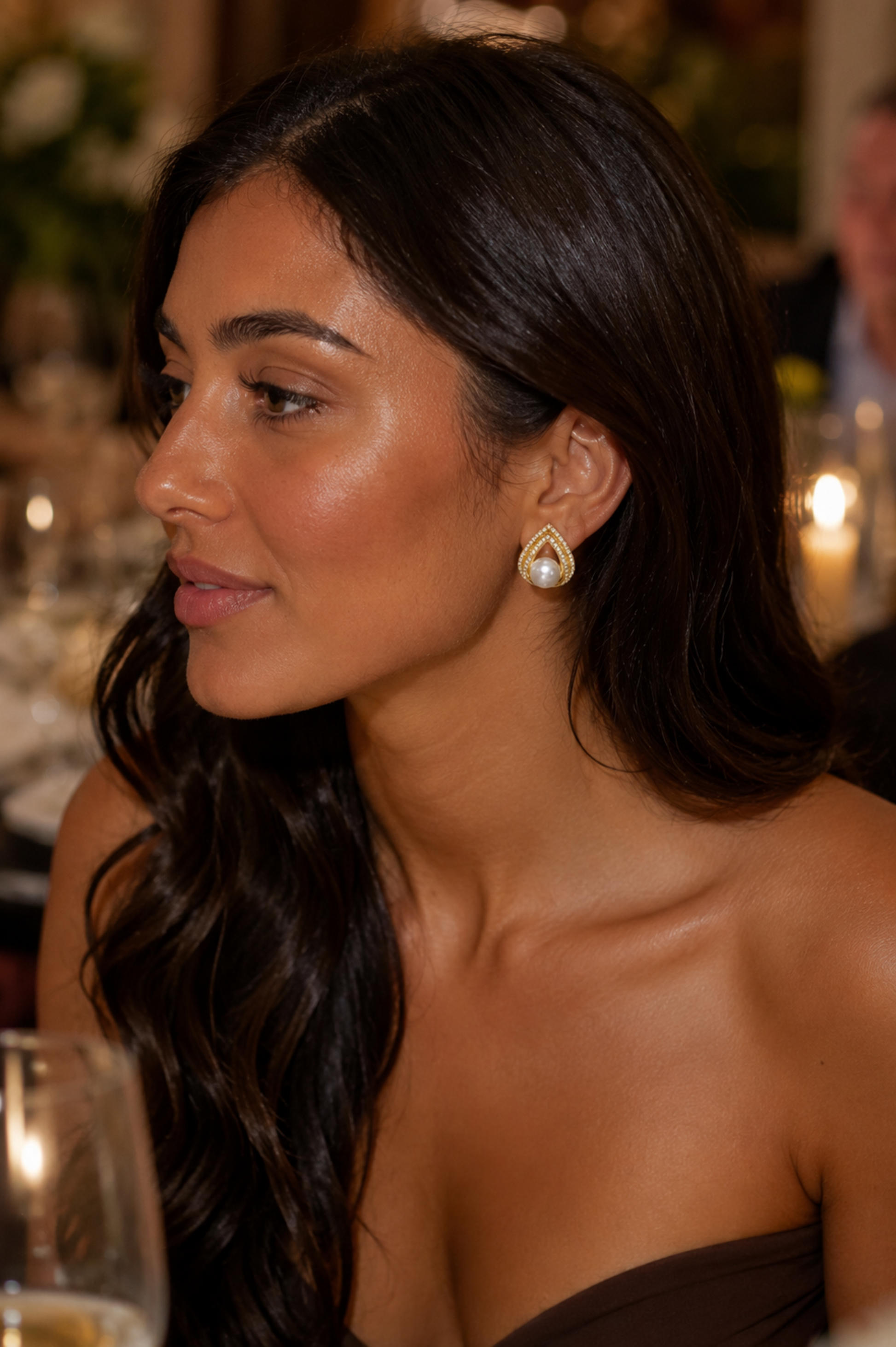 A woman with long dark wavy hair is shown in profile wearing a gold teardrop earring with a white pearl-like center, set against a softly blurred indoor event background.