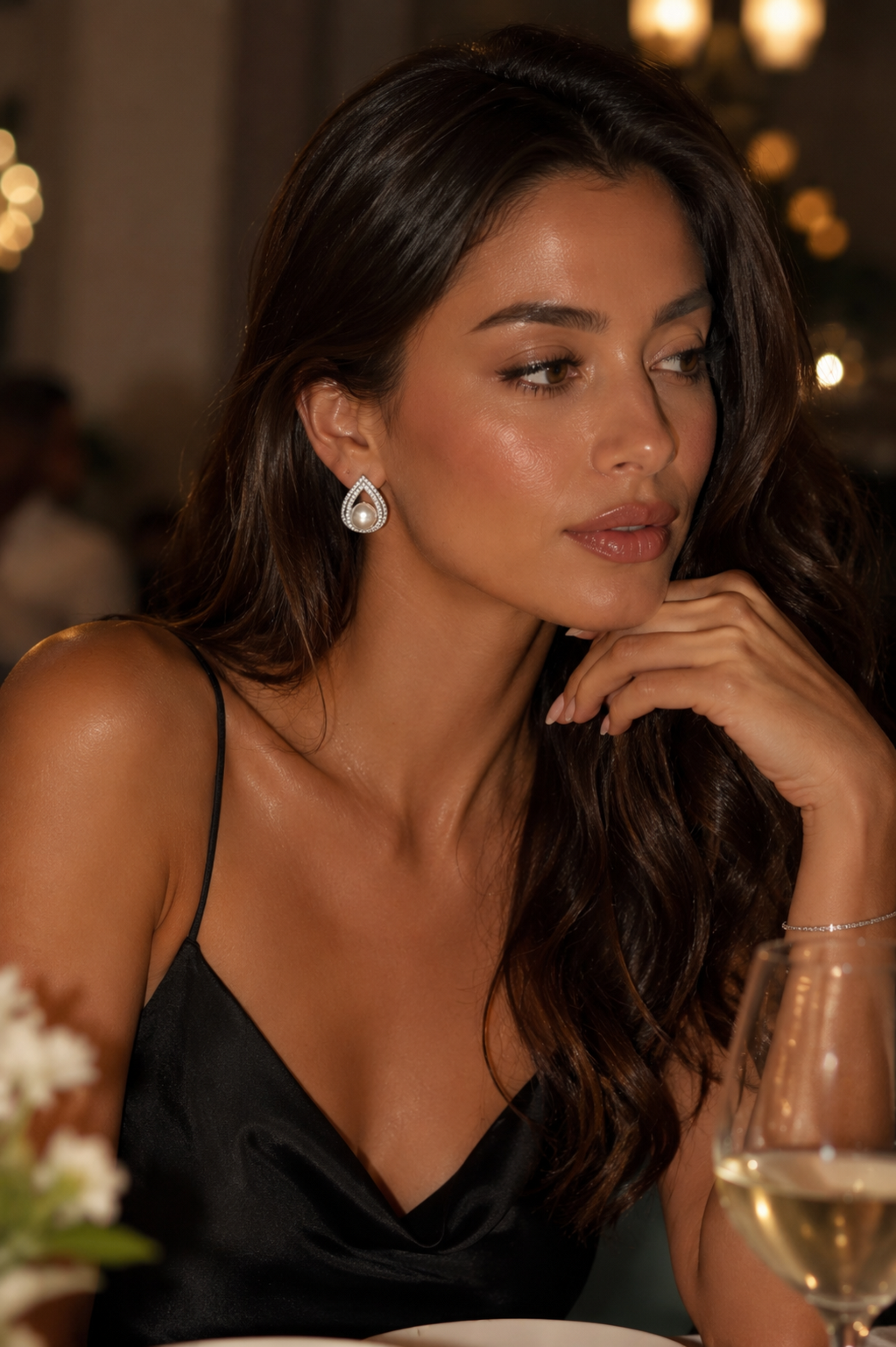 A woman with long dark wavy hair wearing a black satin camisole and a teardrop-shaped silver and white dangle earring, posing at a dinner table with a glass of white wine in the foreground.
