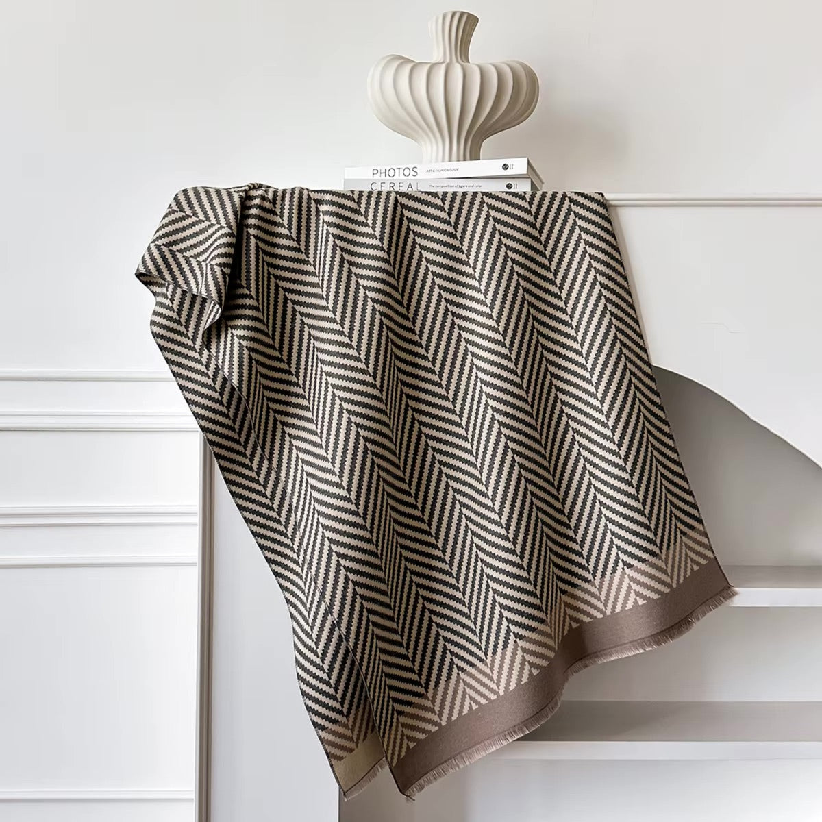 A woven throw blanket with a brown and beige herringbone pattern draped over a white mantel, styled with a decorative ceramic vase and a book.