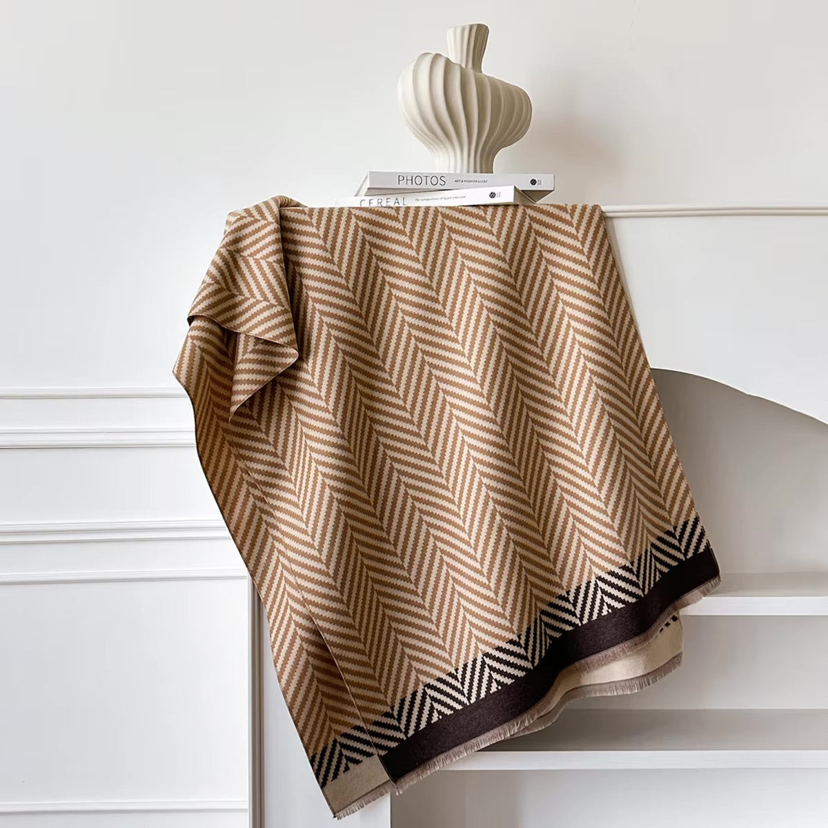 Beige and brown herringbone patterned blanket draped over a white surface with a neutral vase in the background.