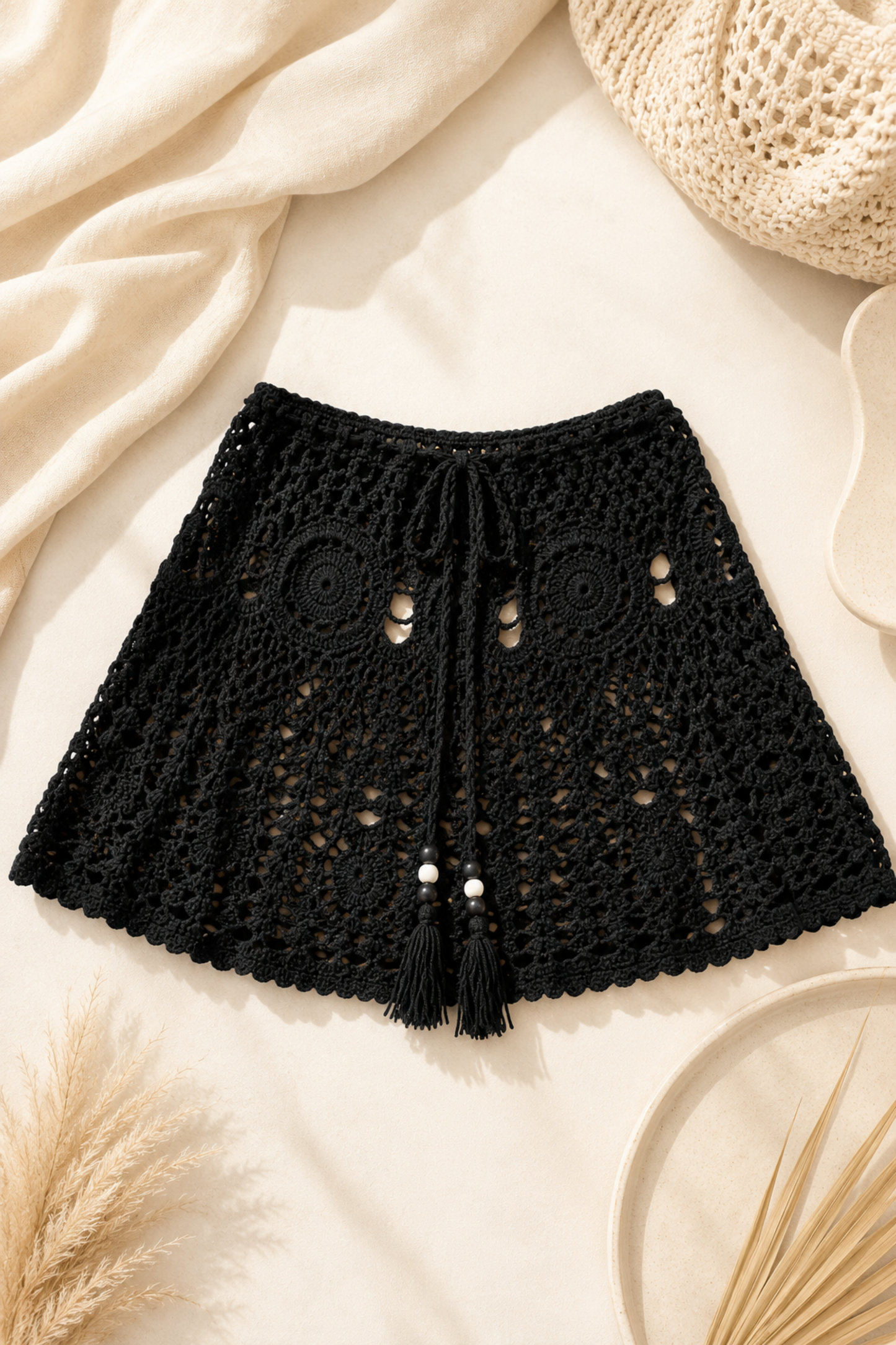 Black crochet knit shorts with a drawstring and tassel, laid flat on a cream fabric background with neutral decorative props.
