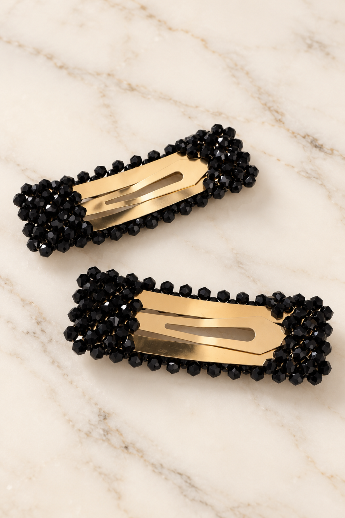 Two black beaded hair clips with gold metal centers, arranged on a light beige marble surface.