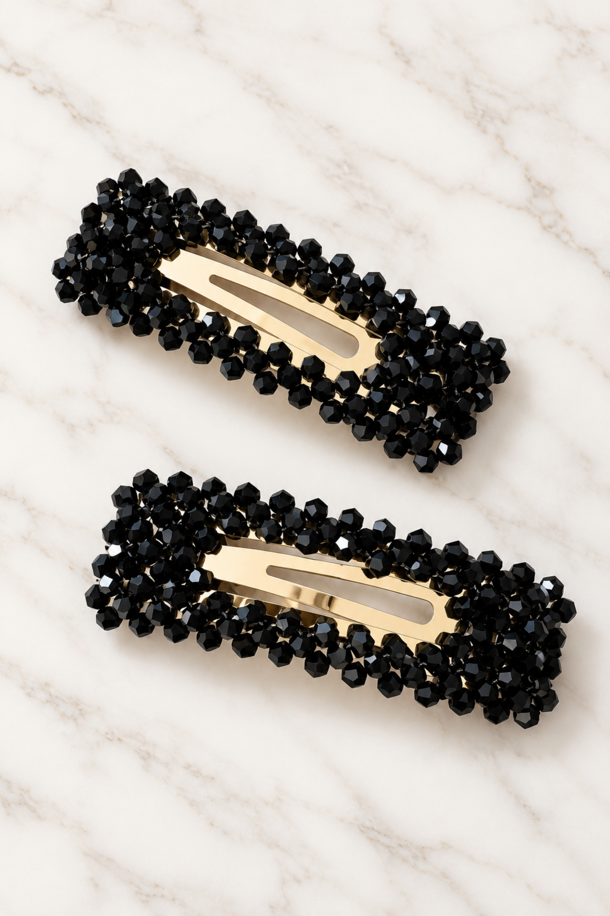 Two black beaded hair clips with gold-tone metal centers, arranged diagonally on a white marble surface.