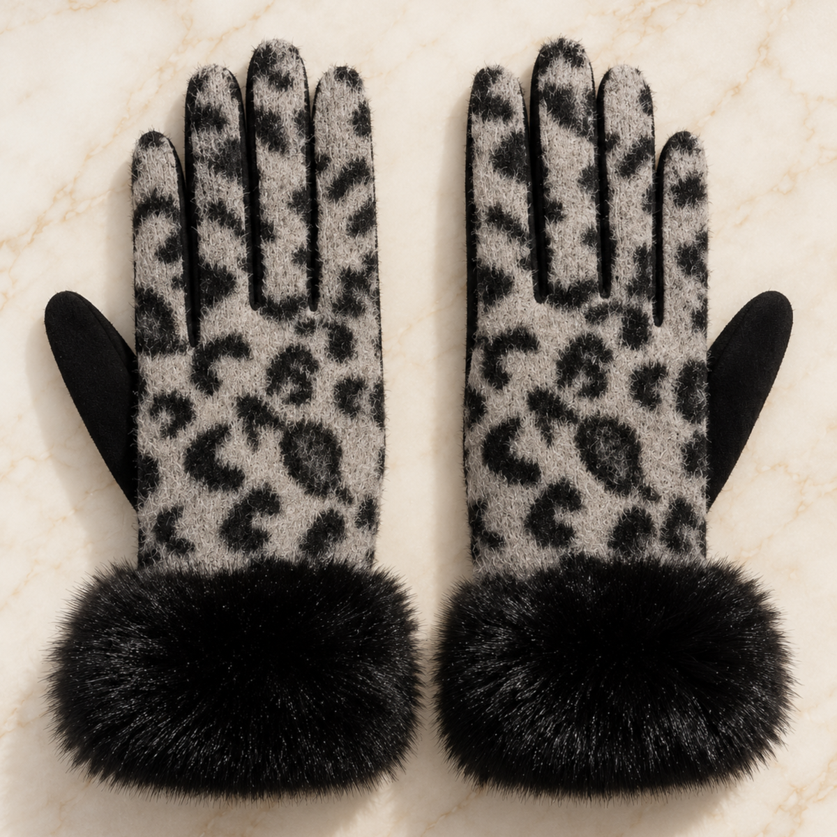 A pair of knit gloves with a black and gray leopard print pattern, featuring solid black fingertips and thick black faux-fur cuffs, displayed on a light beige marble surface.