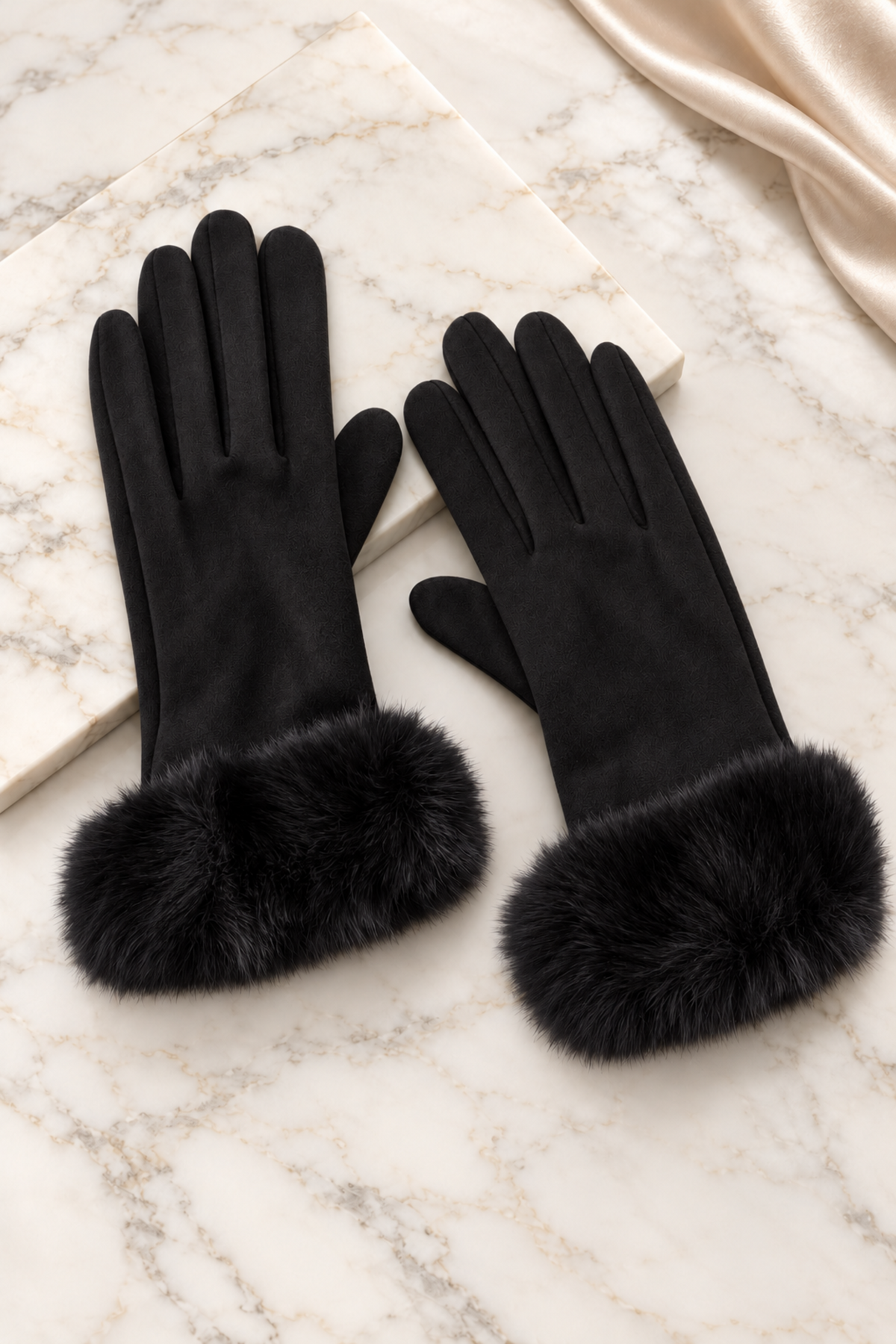 A pair of black leather gloves with plush black fur cuffs, displayed on a white marble surface with a cream satin fabric in the corner.