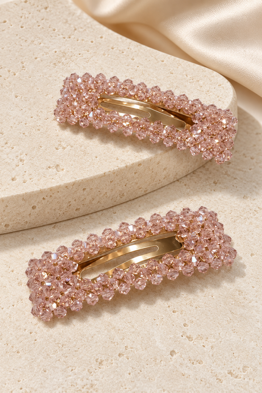 Two rectangular hair clips decorated with rows of pink faceted rhinestones and gold-tone metal, displayed on a textured beige stone surface with a cream satin fabric in the background.