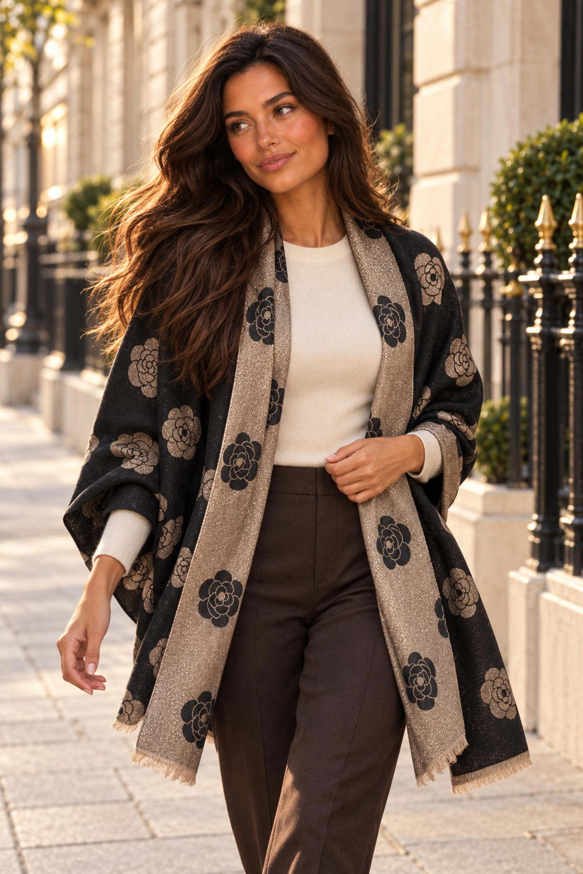 A woman walking outdoors on a city sidewalk wearing a black and beige patterned shawl over a cream top and brown trousers.