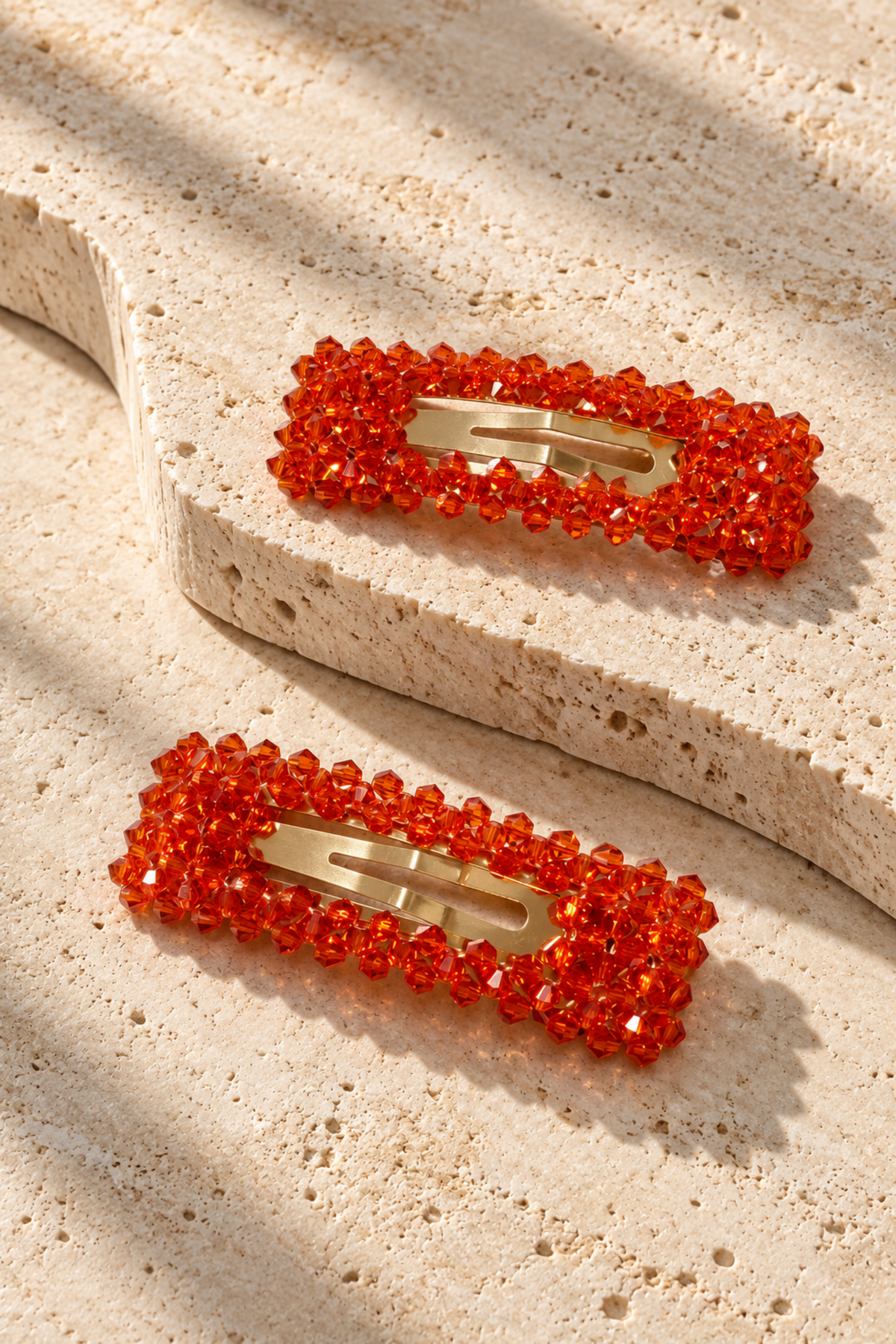 Two rectangular hair clips decorated with bright orange-red faceted beads and gold-tone metal clasps, displayed on a textured beige stone surface with soft shadows.