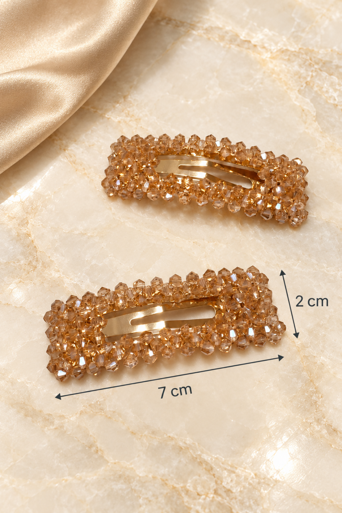 Two rectangular hair clips embellished with faceted champagne-colored rhinestones, displayed on a beige marble surface with a gold satin fabric in the corner. Black lines and text indicate the dimensions of the lower clip as 7 cm by 2 cm.